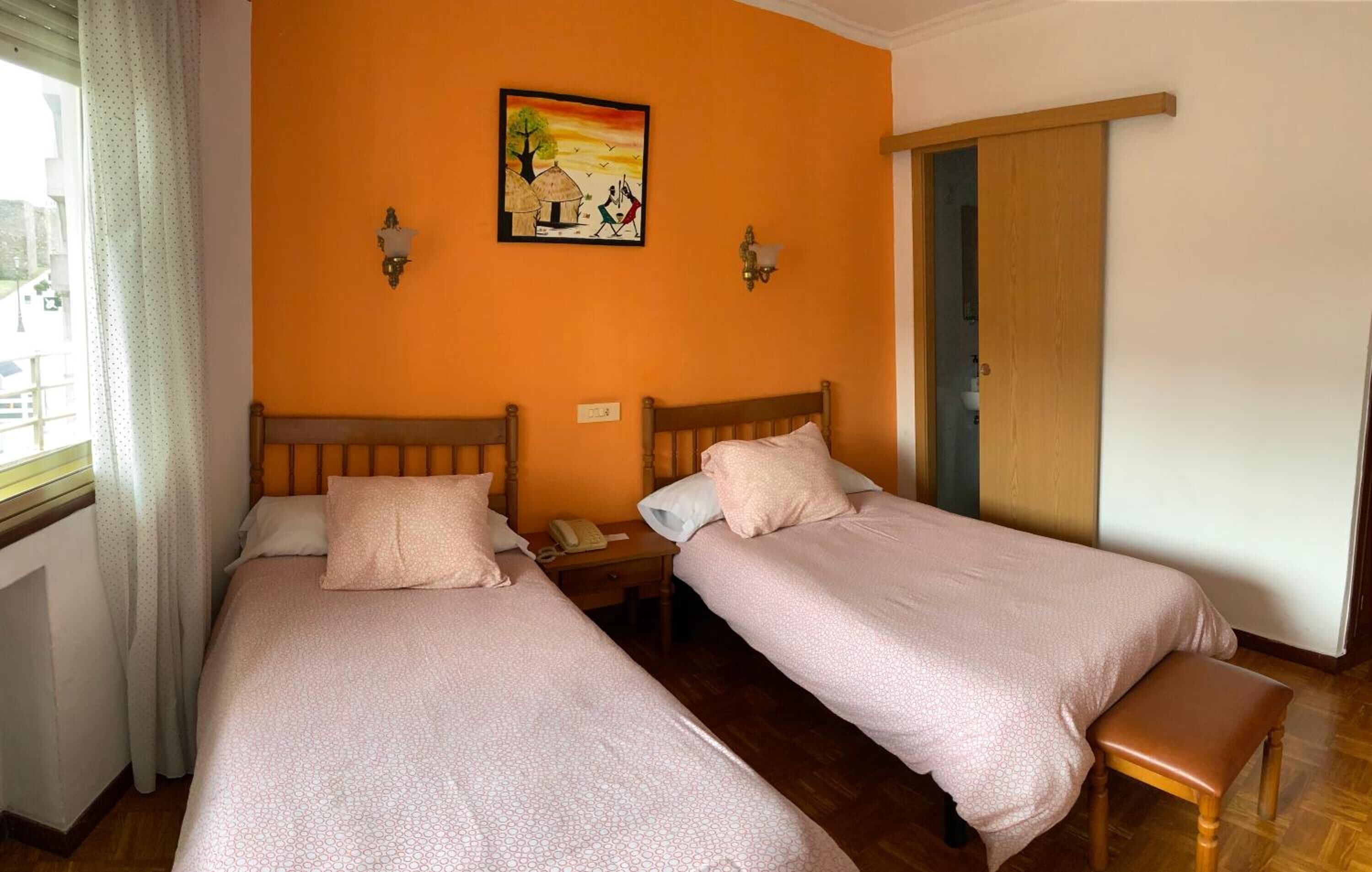 Une chambre avec deux lits, un banc en bois, une table de chevet avec un téléphone et un tableau au mur.
