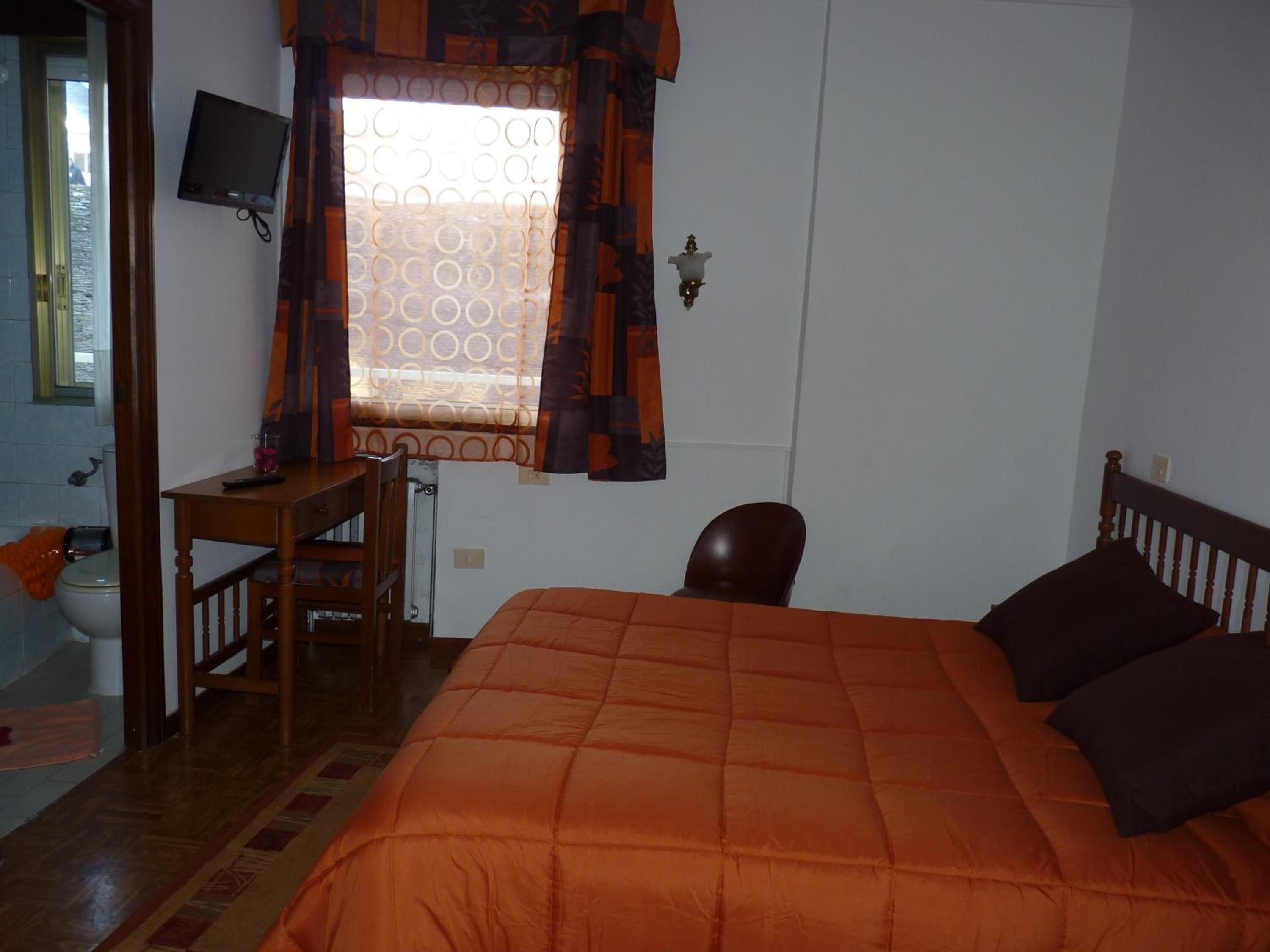 A bedroom with a bed, a desk, a chair, a television, and a window with curtains.