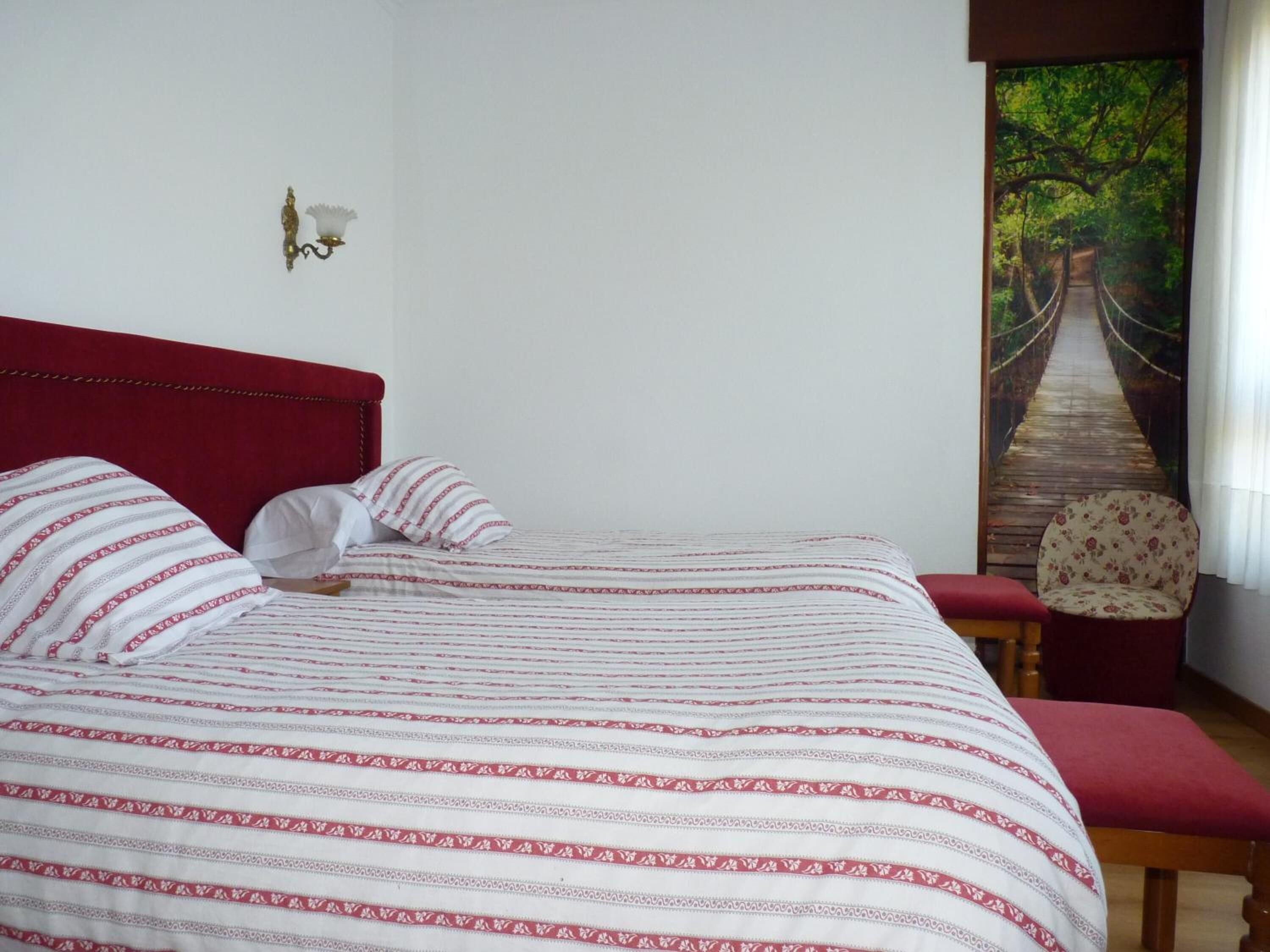 Un dormitorio con un cabecero tapizado rojo, una cama con ropa de cama a rayas, una silla y un cuadro de un puente en un bosque.
