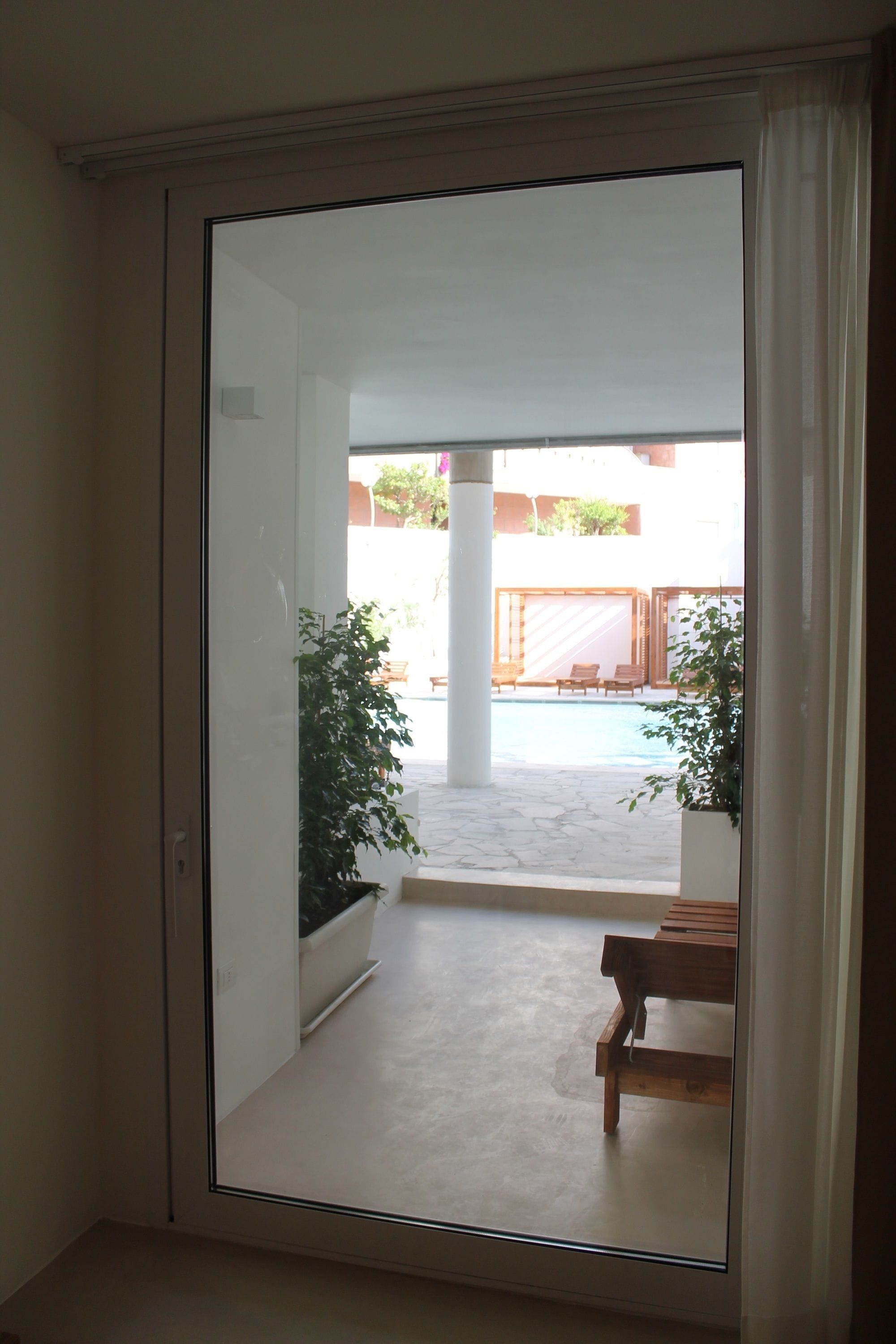 A view through a sliding glass door into an outdoor area with a pool, wooden furniture, and potted plants.