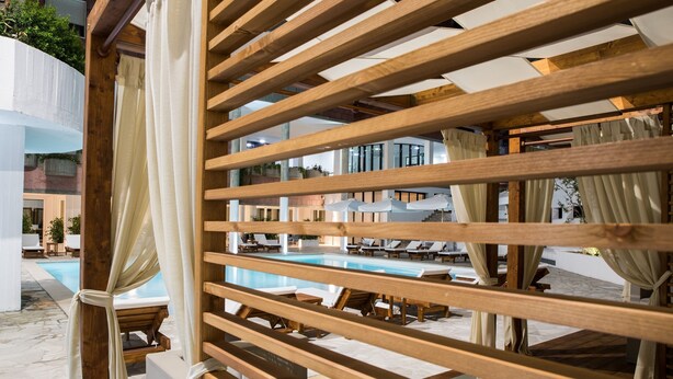A pool area with wooden pergolas and white curtains, surrounded by lounge chairs and umbrellas.