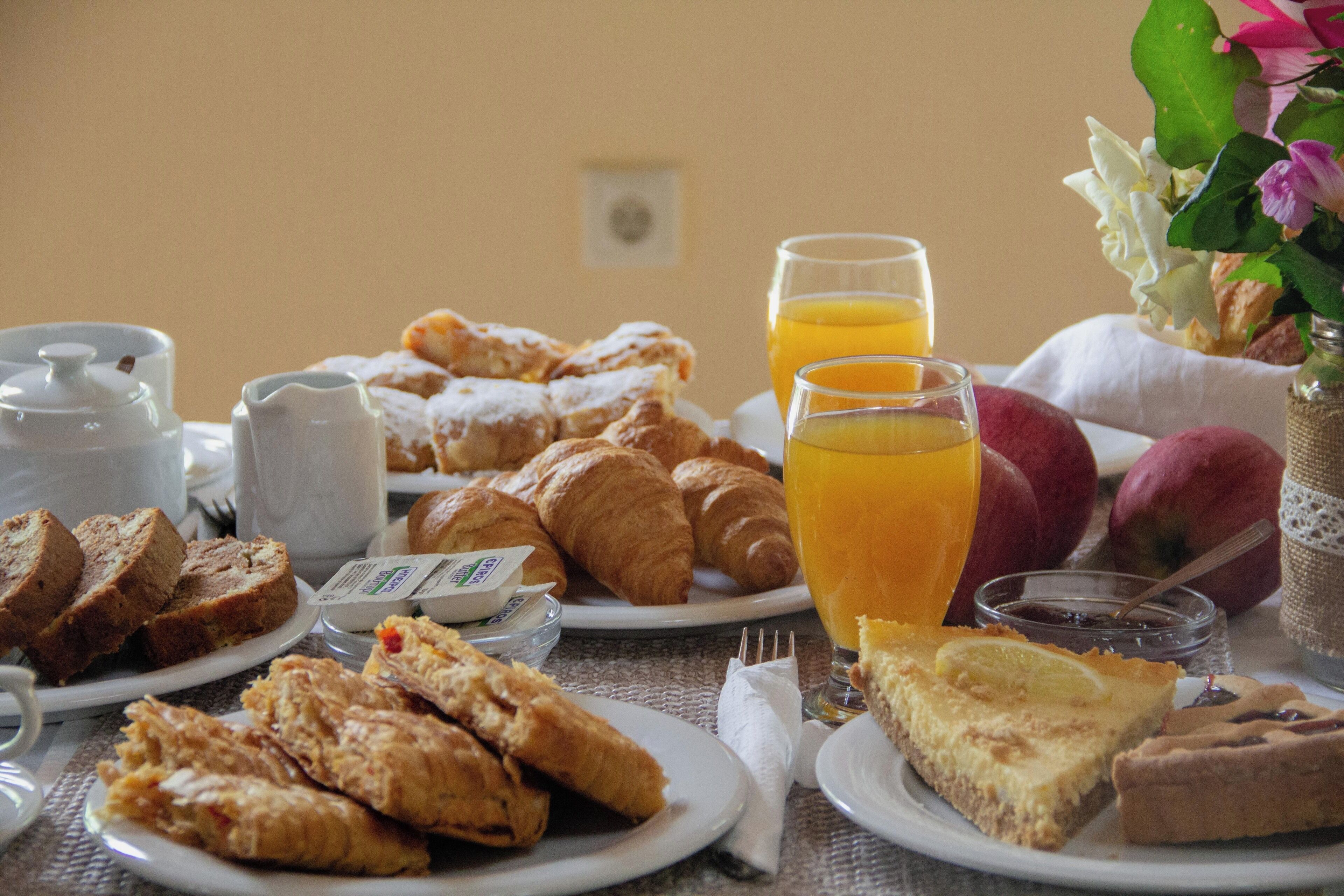 Une table de petit-déjeuner avec des croissants, des viennoiseries, une part de tarte, un verre de jus d’orange et un vase de fleurs.