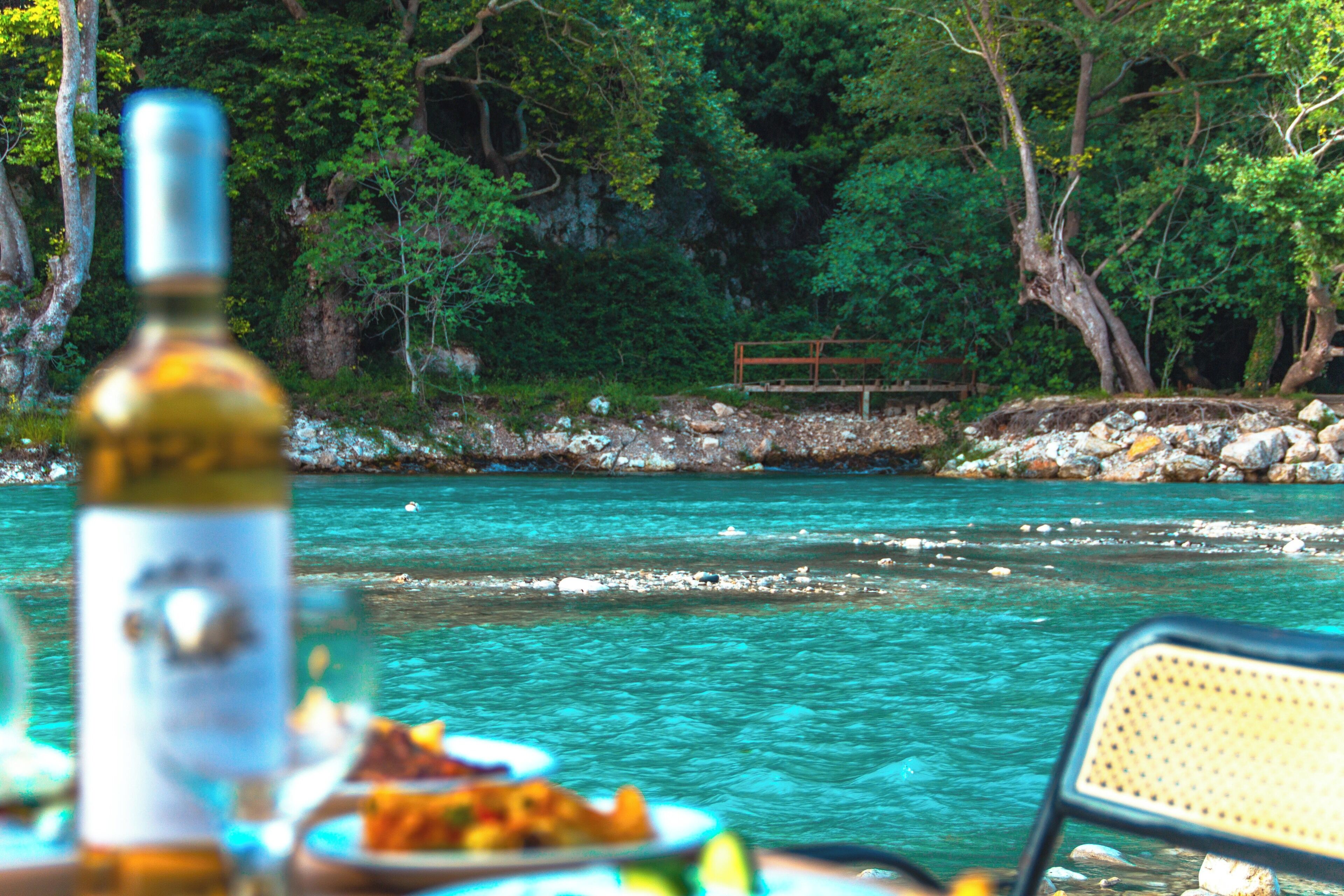 A table with a bottle and food, overlooking a river with rocks and trees in the background.