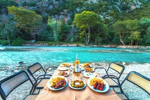 A table set with plates of food and a bottle of wine, overlooking a turquoise river with rocky banks and trees in the background. - Piges Hotel at Acheron Springs (Parga)