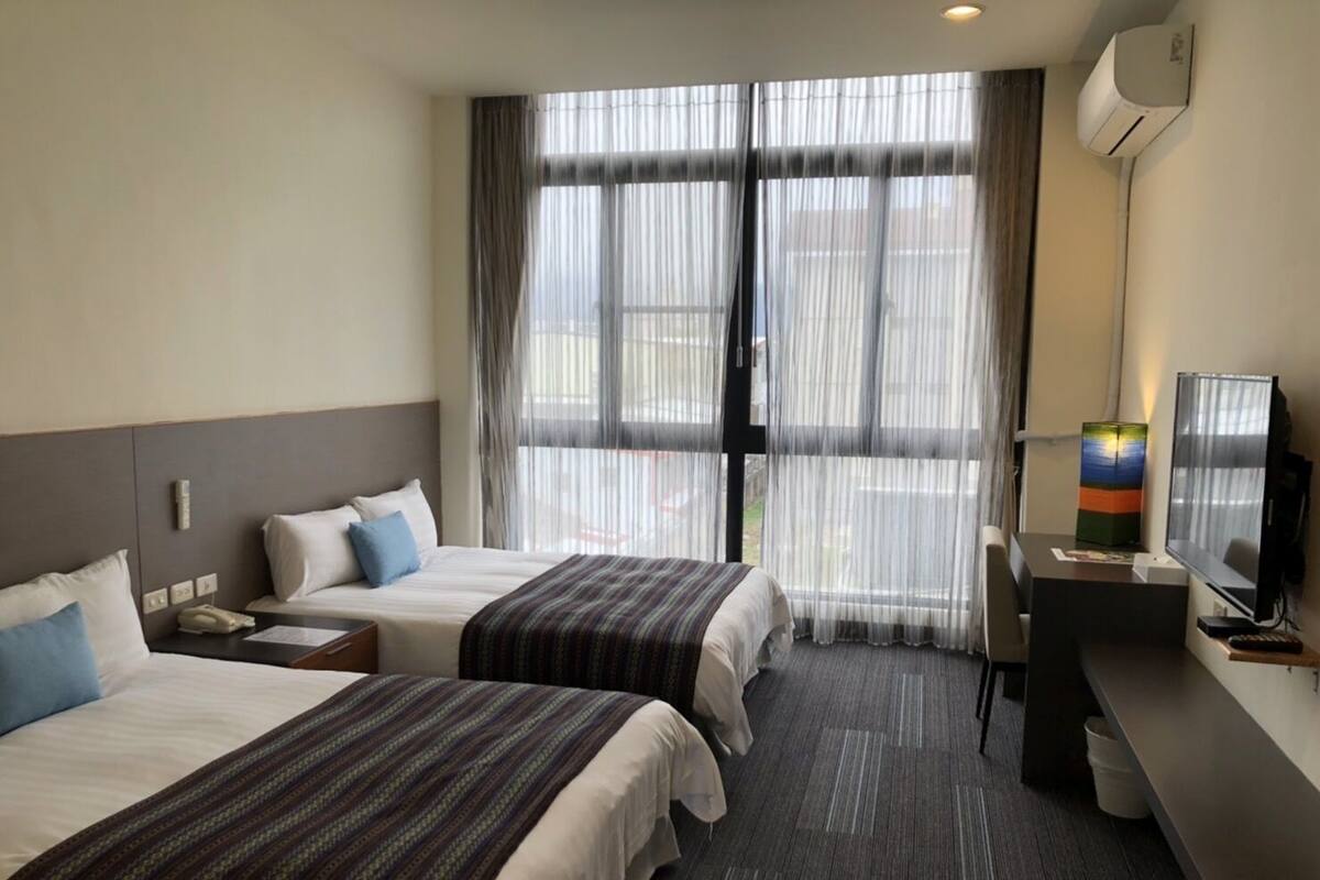 Habitació d'hotel amb dos llits, una taula de treball amb un ordinador i una finestra gran amb cortines translúcides.