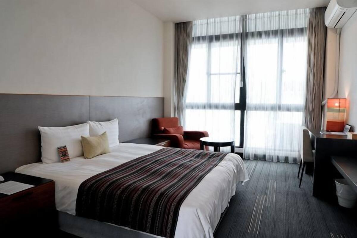 Una habitació d'hotel amb un llit, una taula, una cadira i una finestra amb cortines.