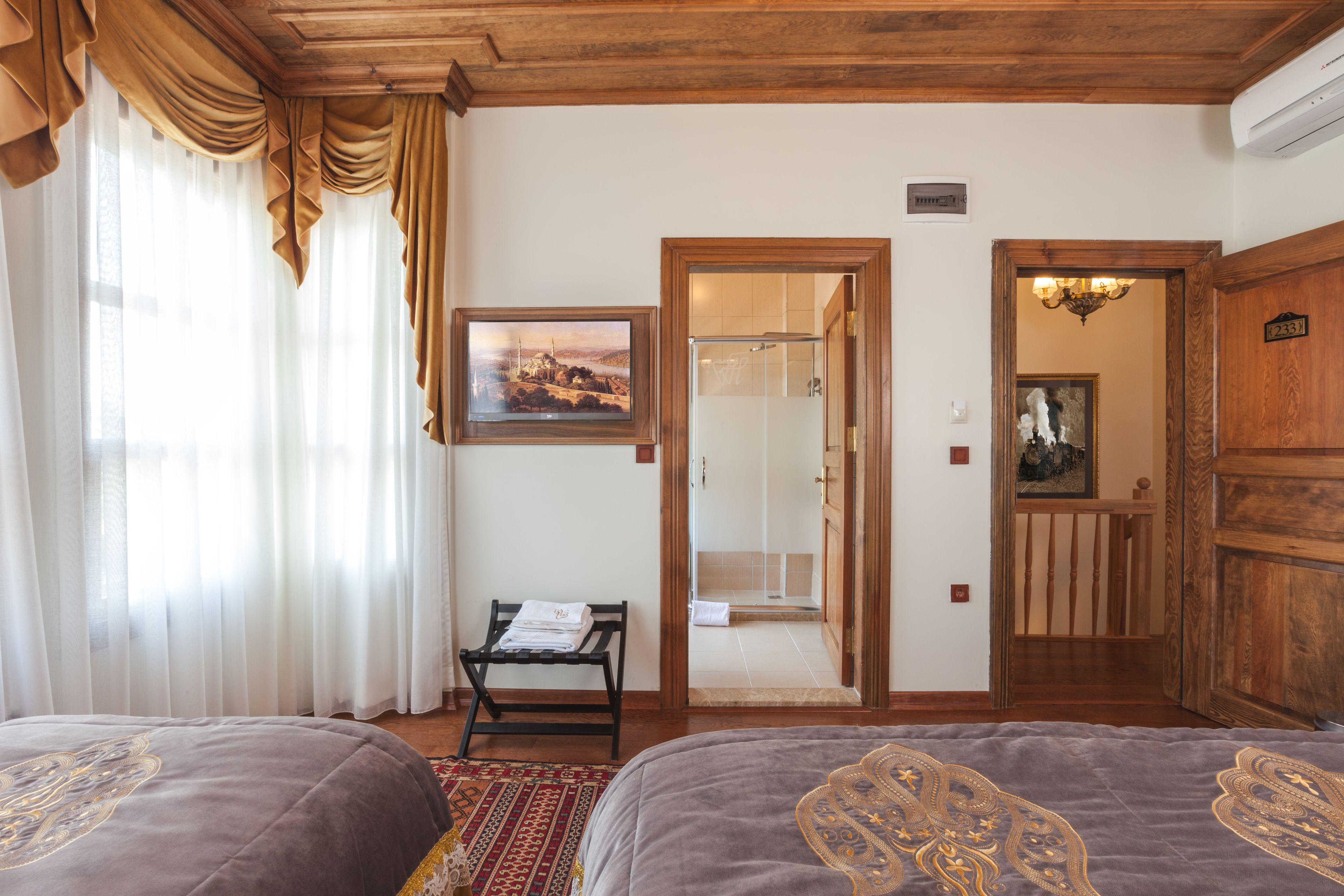 A hotel room with two beds, a wooden ceiling, a framed picture on the wall, a bench with towels, and a door leading to a bathroom.