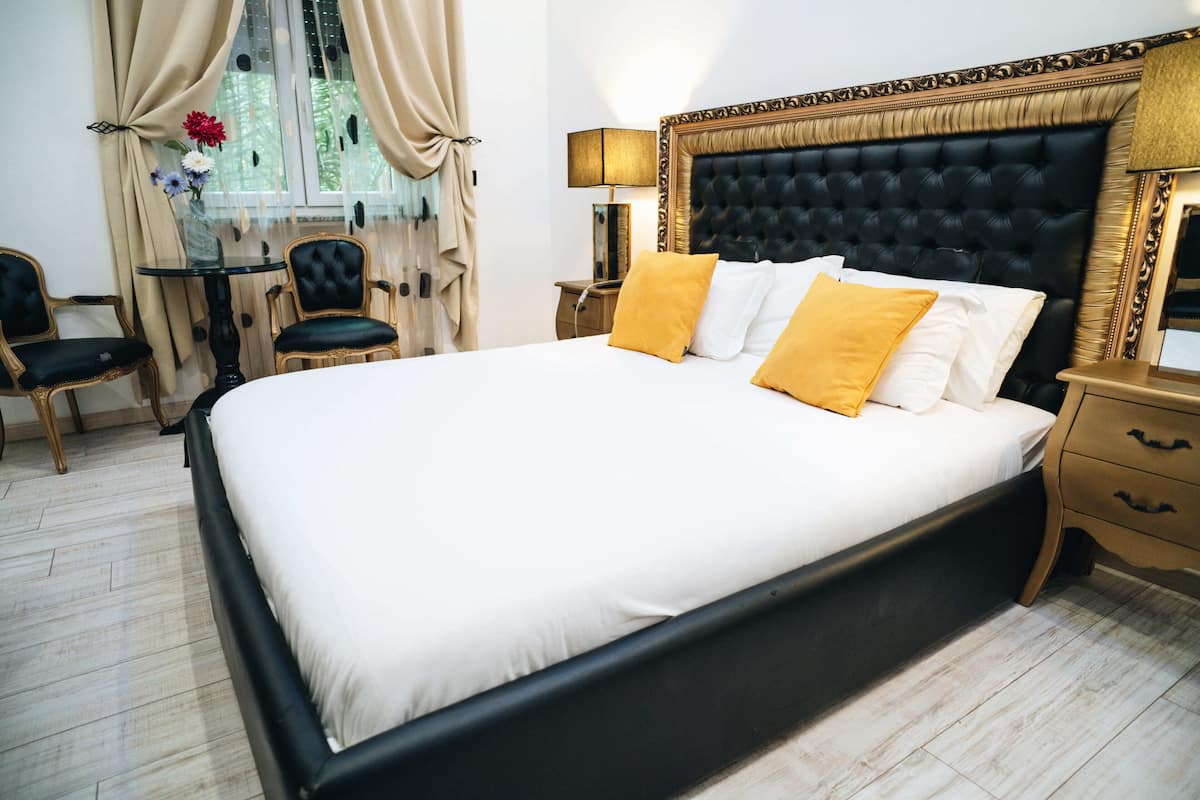Ein ordentlich gemachtes Bett mit schwarzem Kopfteil und weißer Bettwäsche, ein kleiner Tisch mit Stühlen und ein Fenster mit Vorhängen.