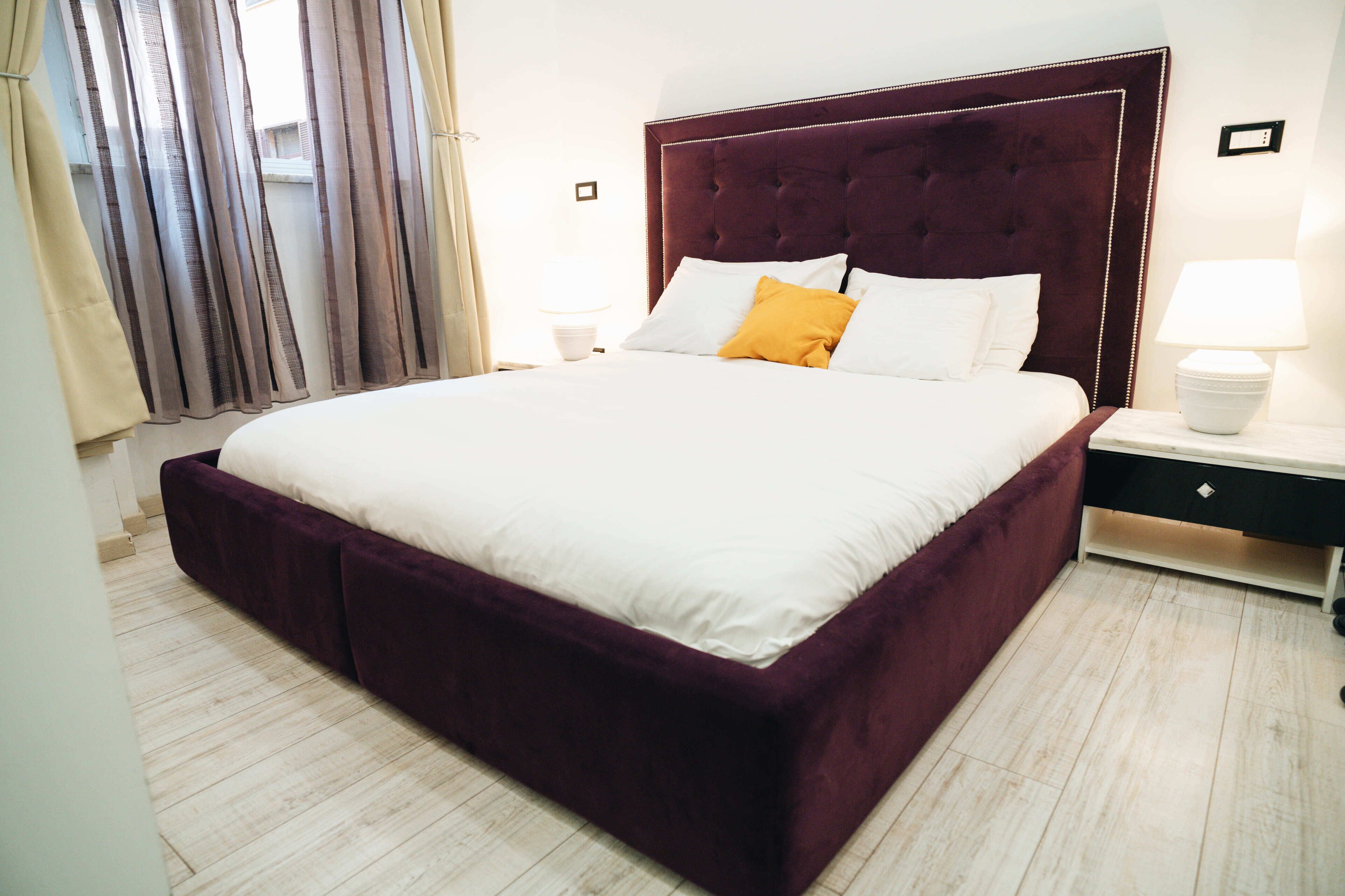 A large bed with a dark headboard, white bedding, and a mustard-colored pillow. There is a bedside table with a lamp and a small drawer. The room has light wooden flooring and a window with curtains.