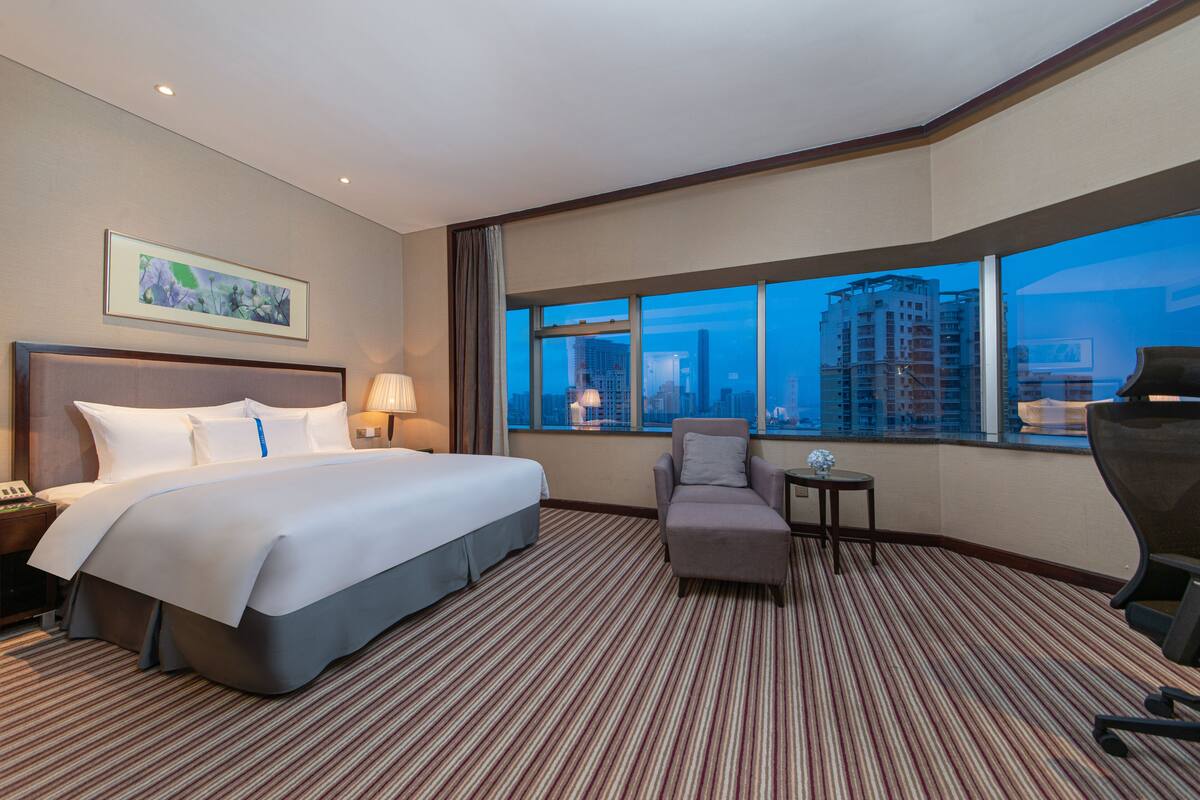 Hotelkamer met een groot bed, een stoel, een kleine tafel en uitzicht op de stad door grote ramen.