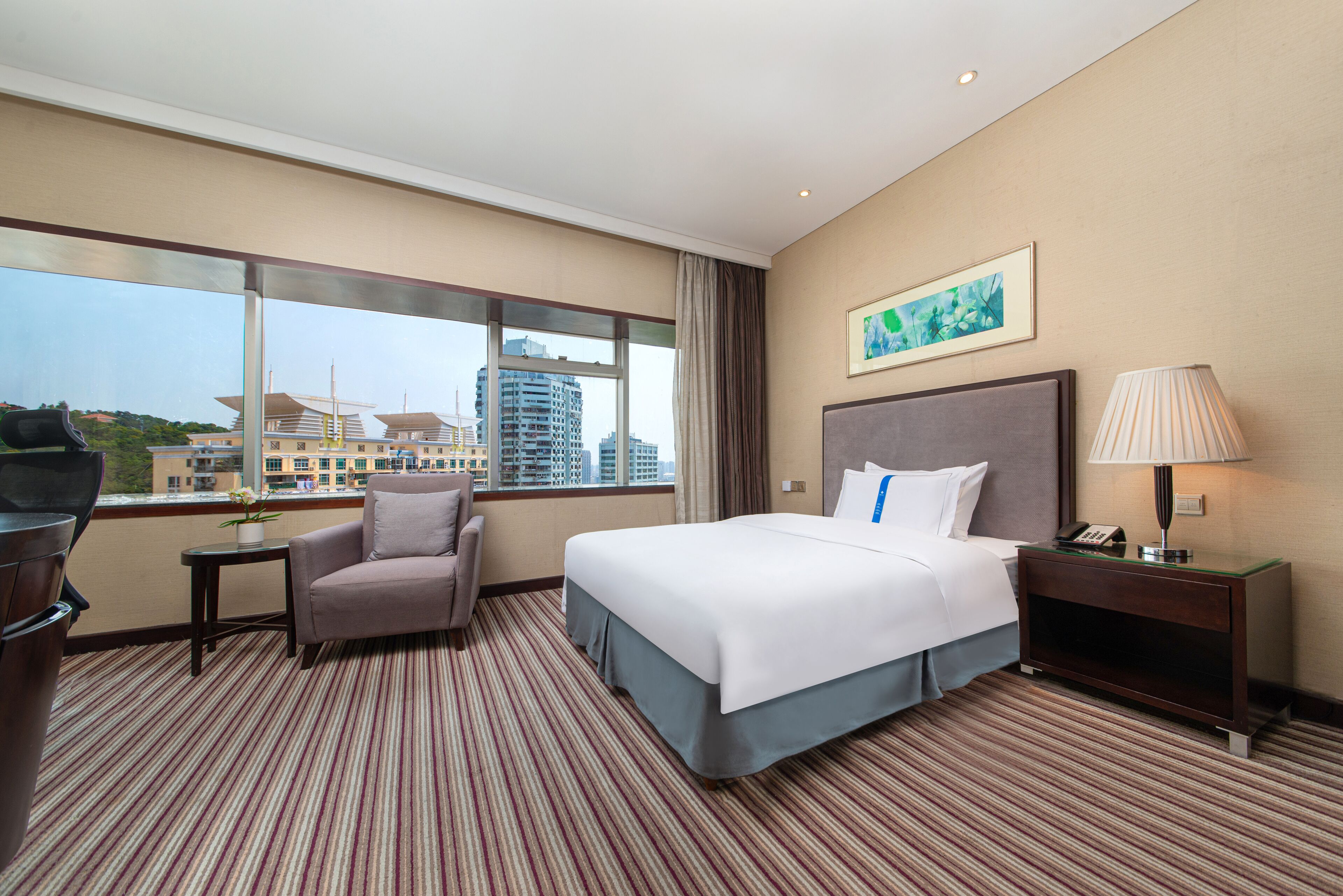 Quarto de hotel com uma cama grande, uma cadeira, uma mesinha, uma mesa de cabeceira e vista para a cidade através de grandes janelas.