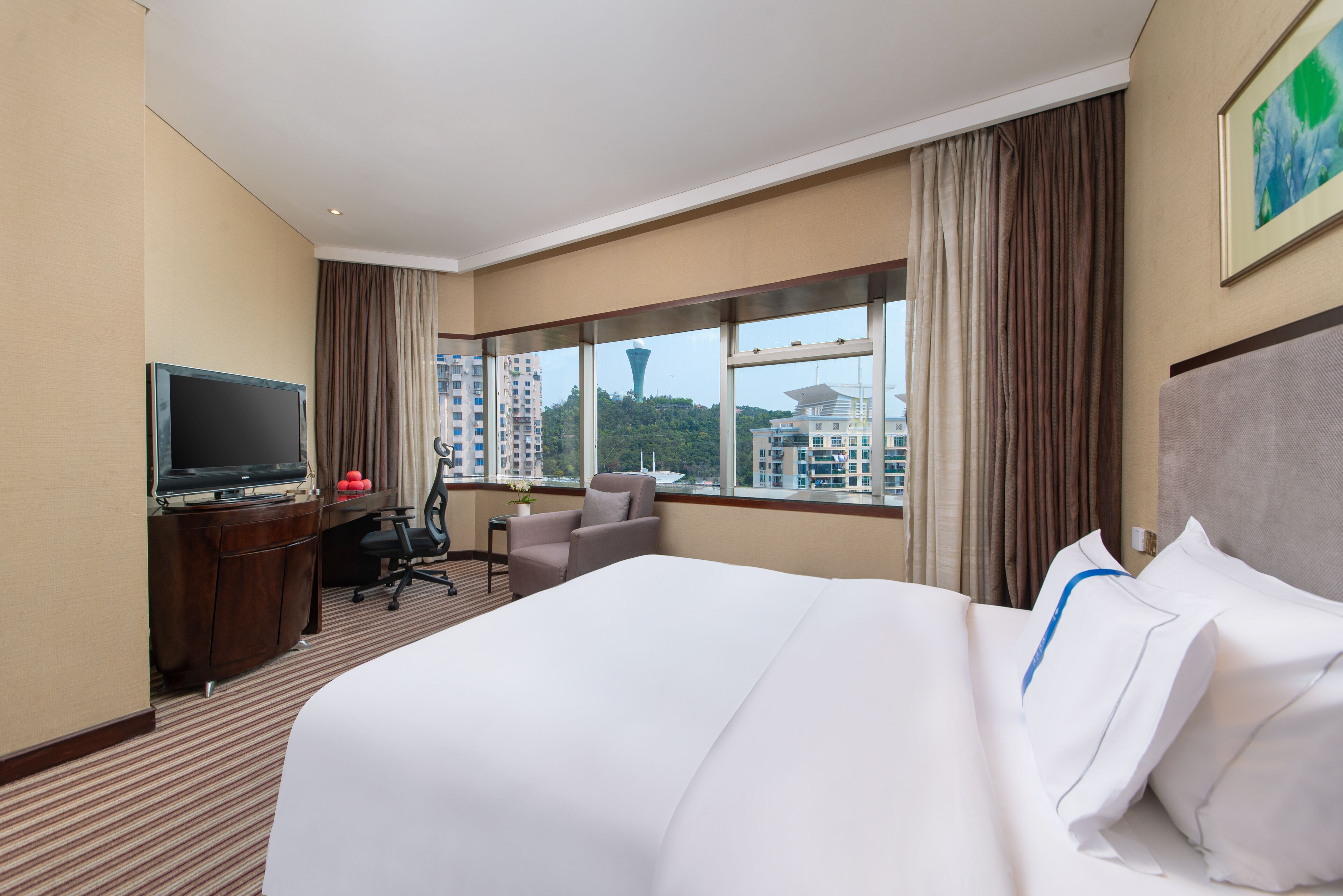 A hotel room with a large bed, a desk with a TV, a chair, and a view of buildings and a tower outside the window.