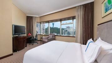 A hotel room with a large bed, a desk with a TV, a chair, and a view of buildings and a tower outside the window.