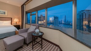Ein Hotelzimmer mit Bett, Sitzecke mit Sessel und Hocker, kleinem Tisch mit Vase und großen Fenstern mit Blick auf die Stadt.