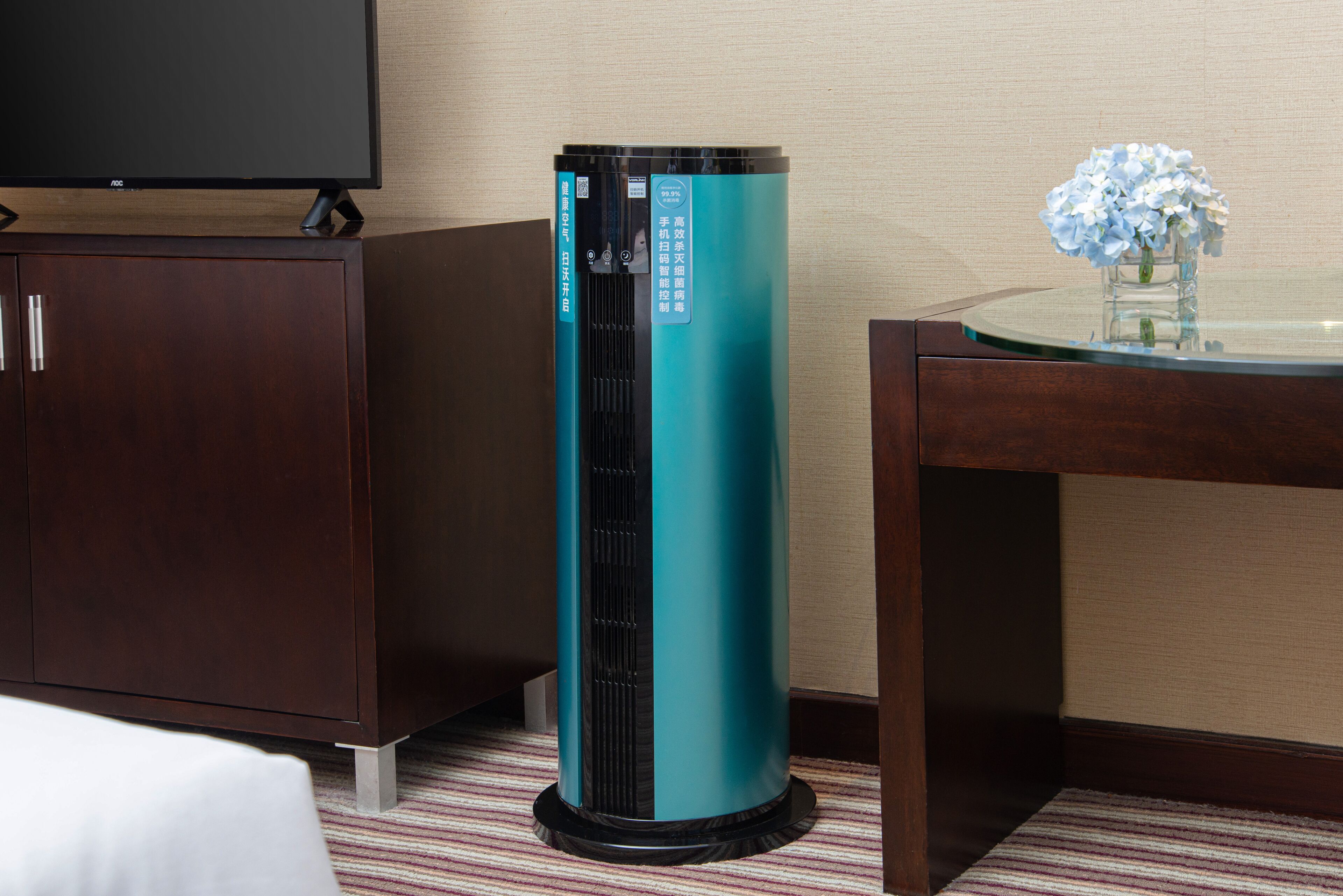 A teal air purifier, a flat-screen TV, a wooden cabinet, and a glass table with a vase of flowers.