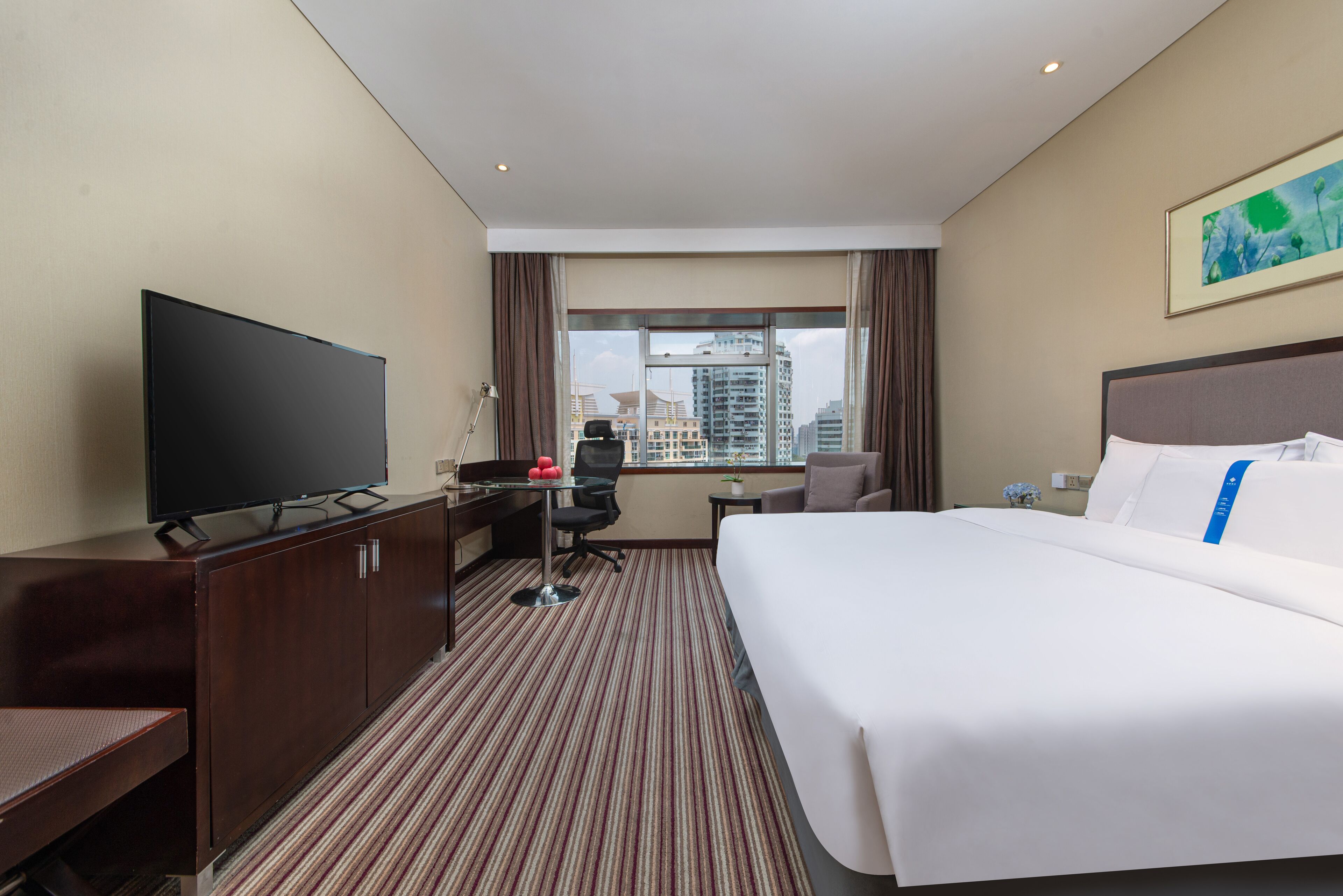 A hotel room with a large bed, a desk with a TV, a chair, and a view of the cityscape through the window.