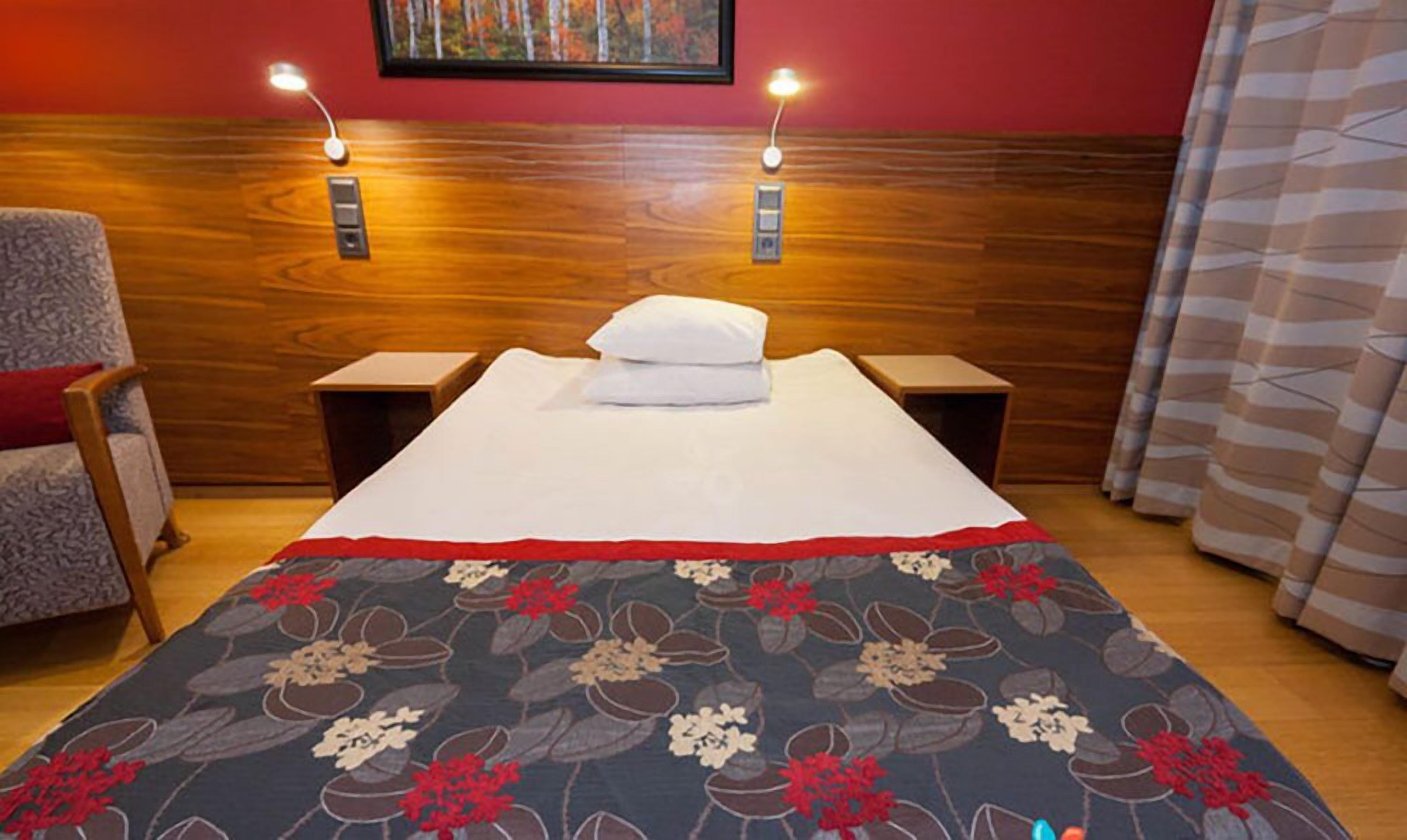 Habitación de hotel con cabecera de madera, dos lámparas de noche, cama con colcha estampada, una silla y un cuadro en la pared.