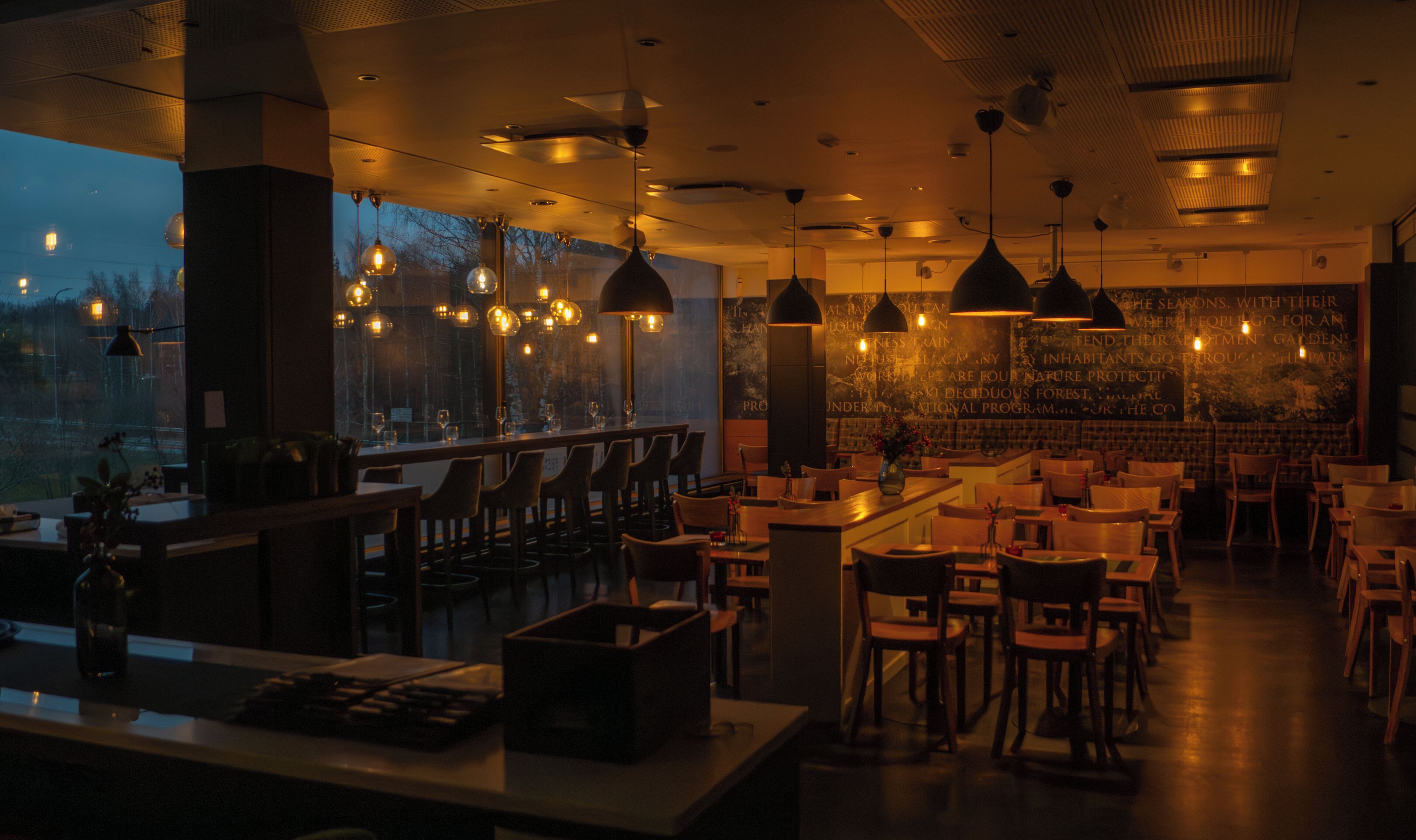Un interior de restaurante con varias mesas y sillas, iluminación colgante y vistas a través de las ventanas.