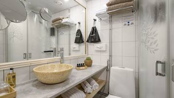 Un baño moderno con un espejo grande, un lavamanos de piedra color beige, ducha e inodoro. Hay toallas y artículos de aseo en la encimera.