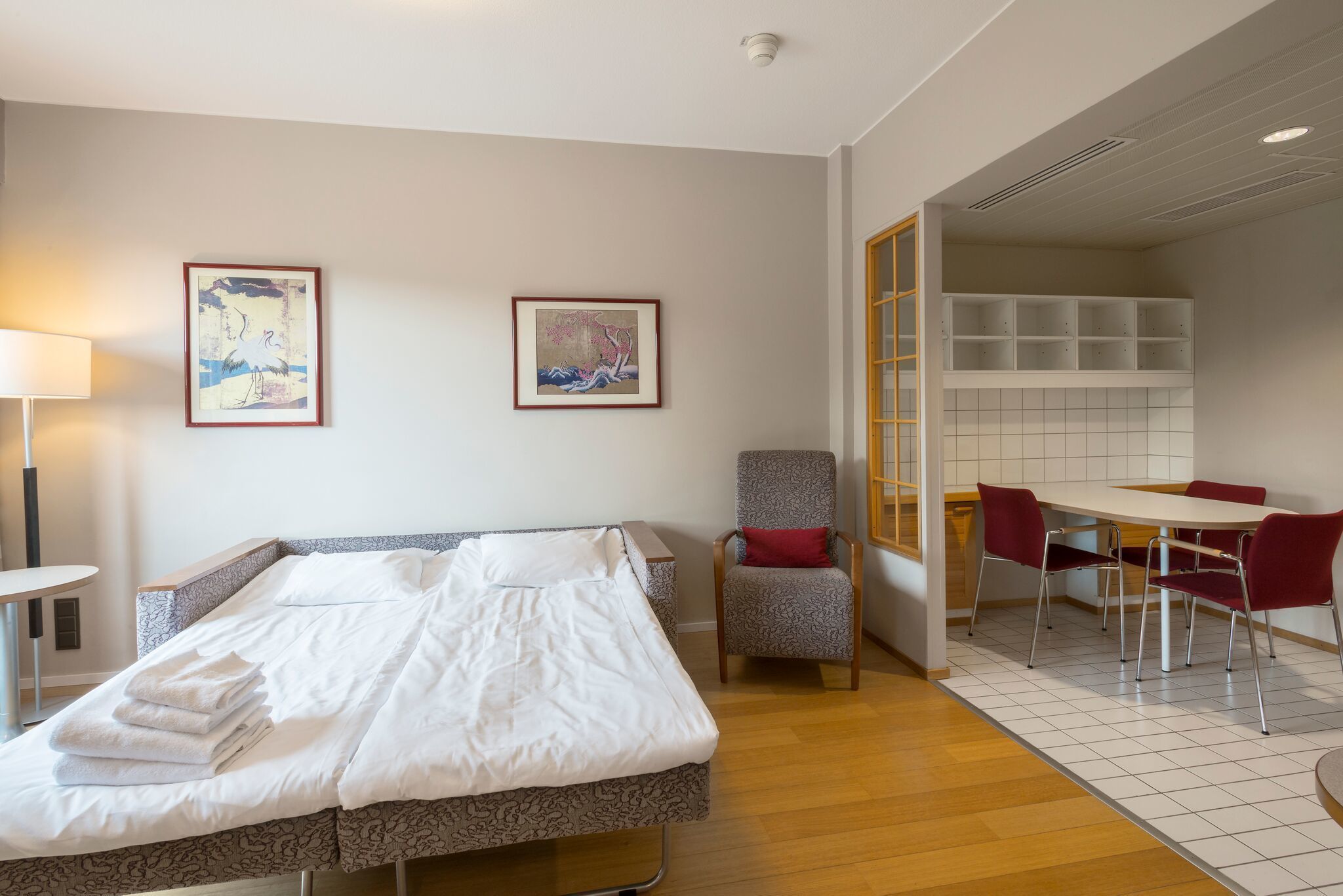 A hotel room with a bed, a chair, a table, and two framed pictures on the wall.