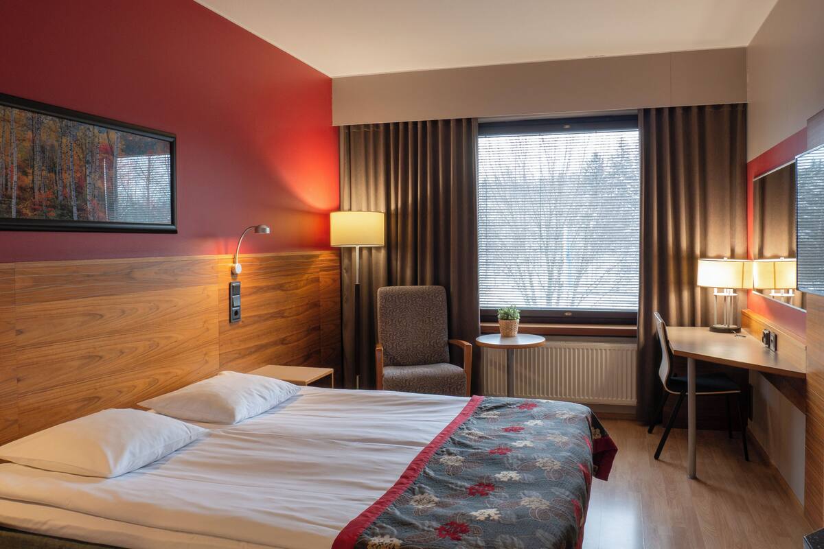 Habitación de hotel con cama, escritorio, silla, lámpara, ventana con cortinas y un cuadro en la pared.