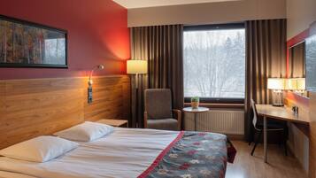 A hotel room with a bed, a desk, a chair, a lamp, a window with curtains, and a painting on the wall.