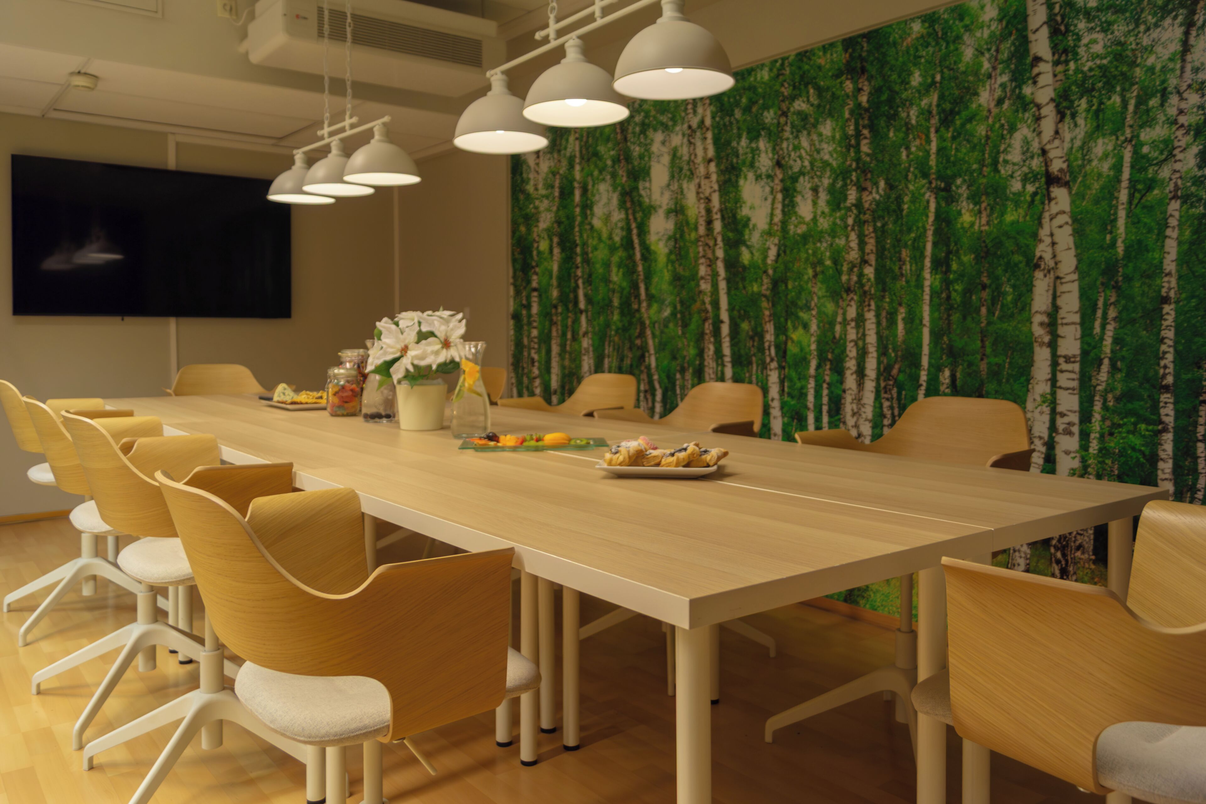 A conference room with a long table, chairs, a TV screen, and a wall with a tree mural.