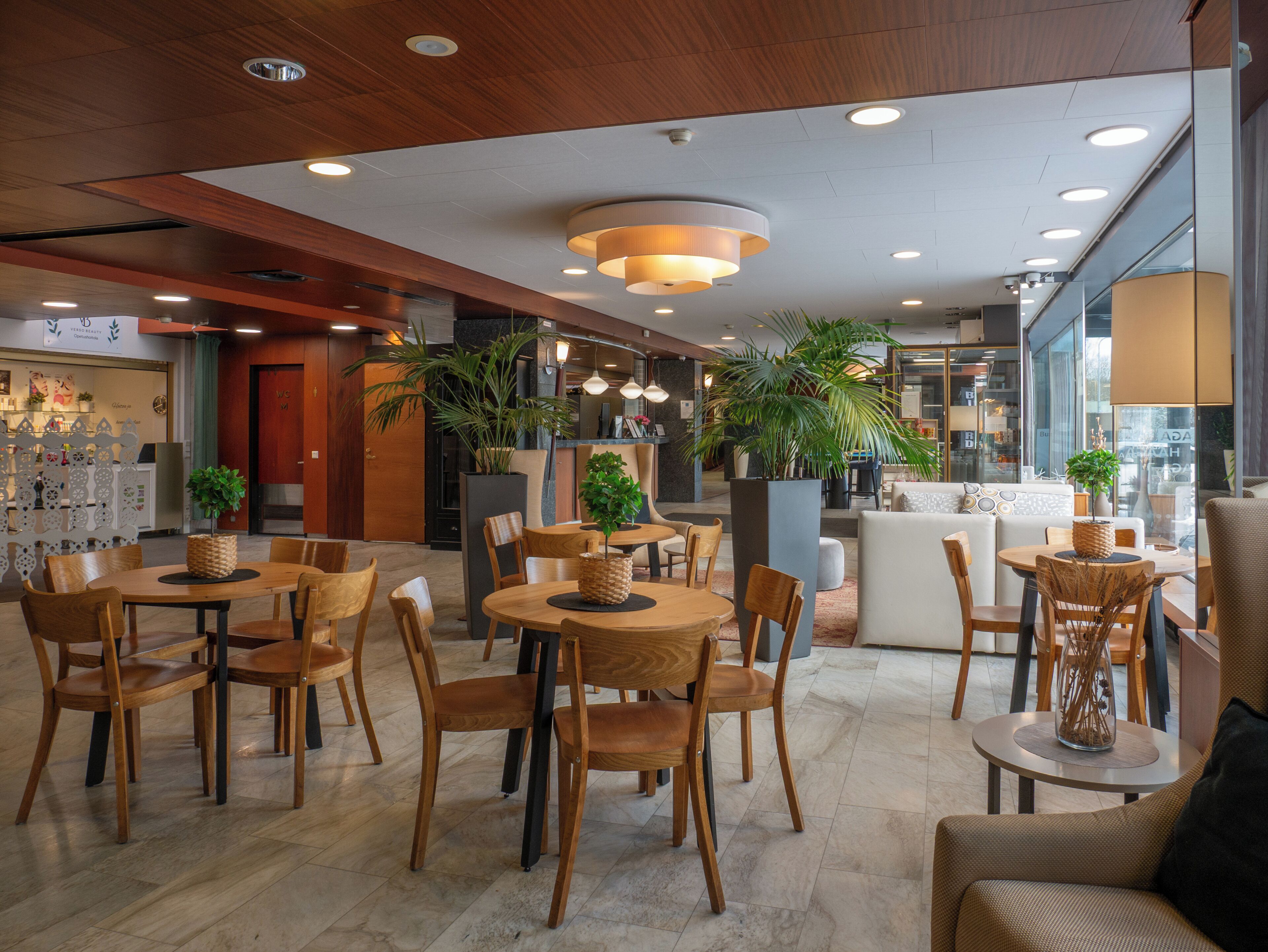 A modern hotel lobby with wooden flooring, round tables, and chairs, a reception desk, and large potted plants.