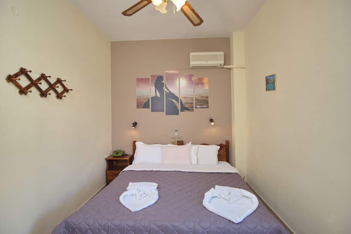A hotel room with a bed, a wall-mounted air conditioner, a bedside table with a lamp, and a wall art piece.