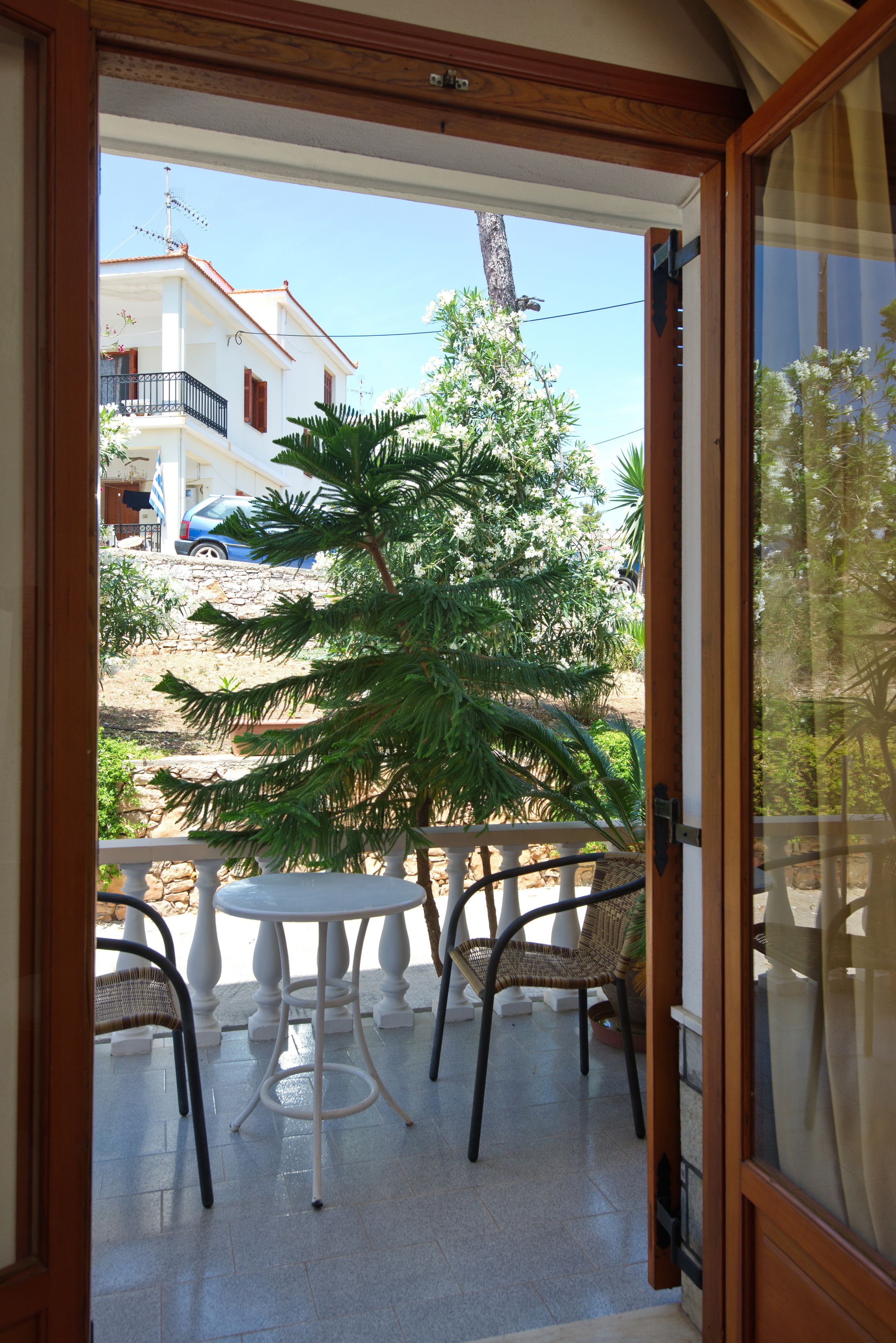 Una puerta abierta que da a un balcón con mesa y sillas, vista a una casa y un árbol.