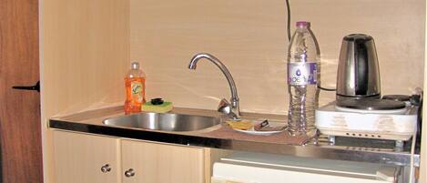 A compact kitchen with a sink, a small refrigerator, a kettle, and a bottle of water.