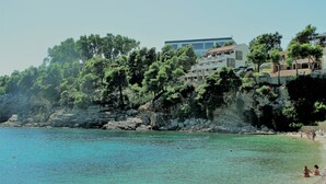 Uma área costeira com águas turquesa cristalinas, falésias rochosas e edifícios com varandas viradas para o mar.