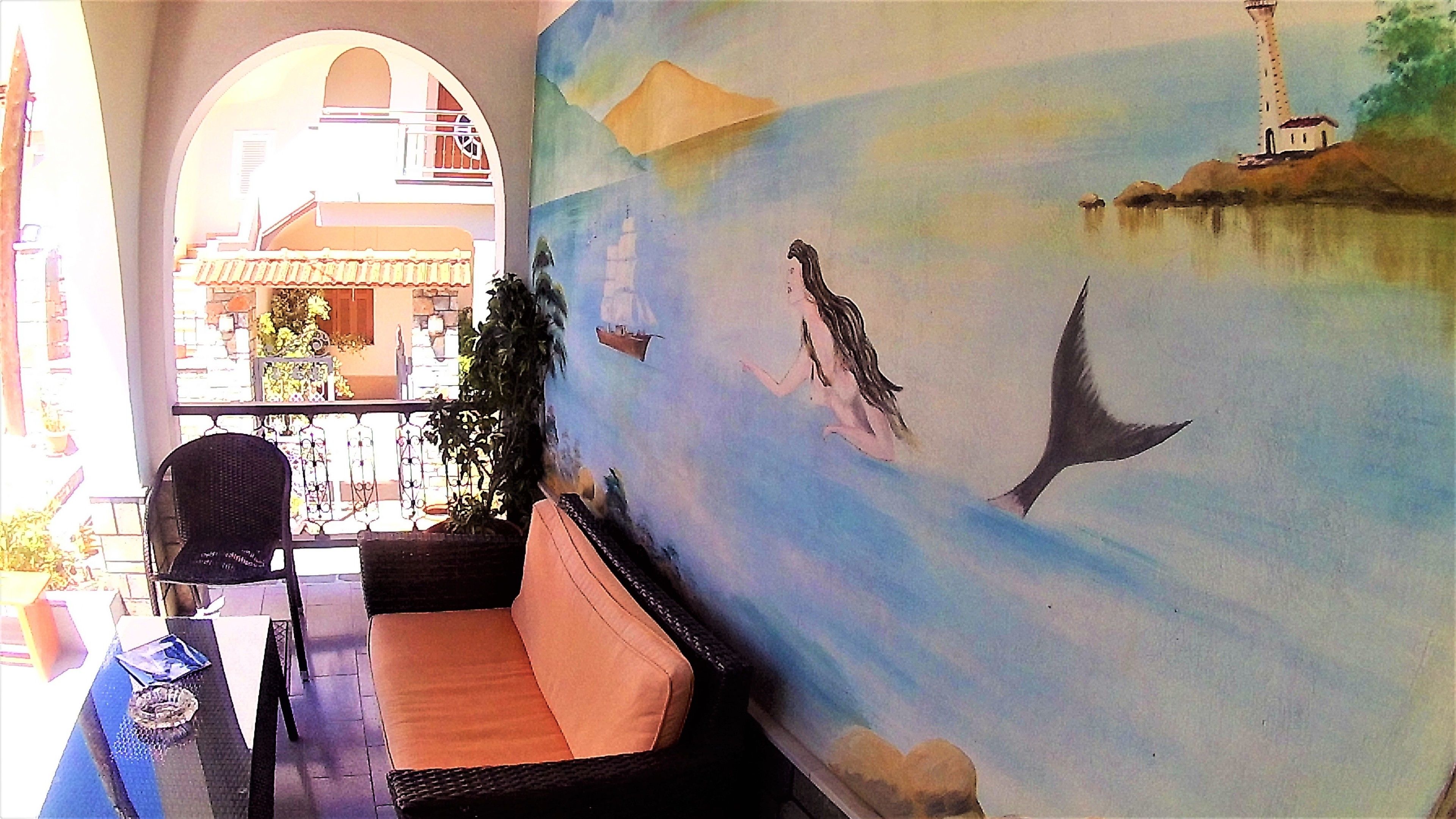 A room with a mural of a mermaid and a lighthouse, a wicker sofa, and a view of a building with a balcony.