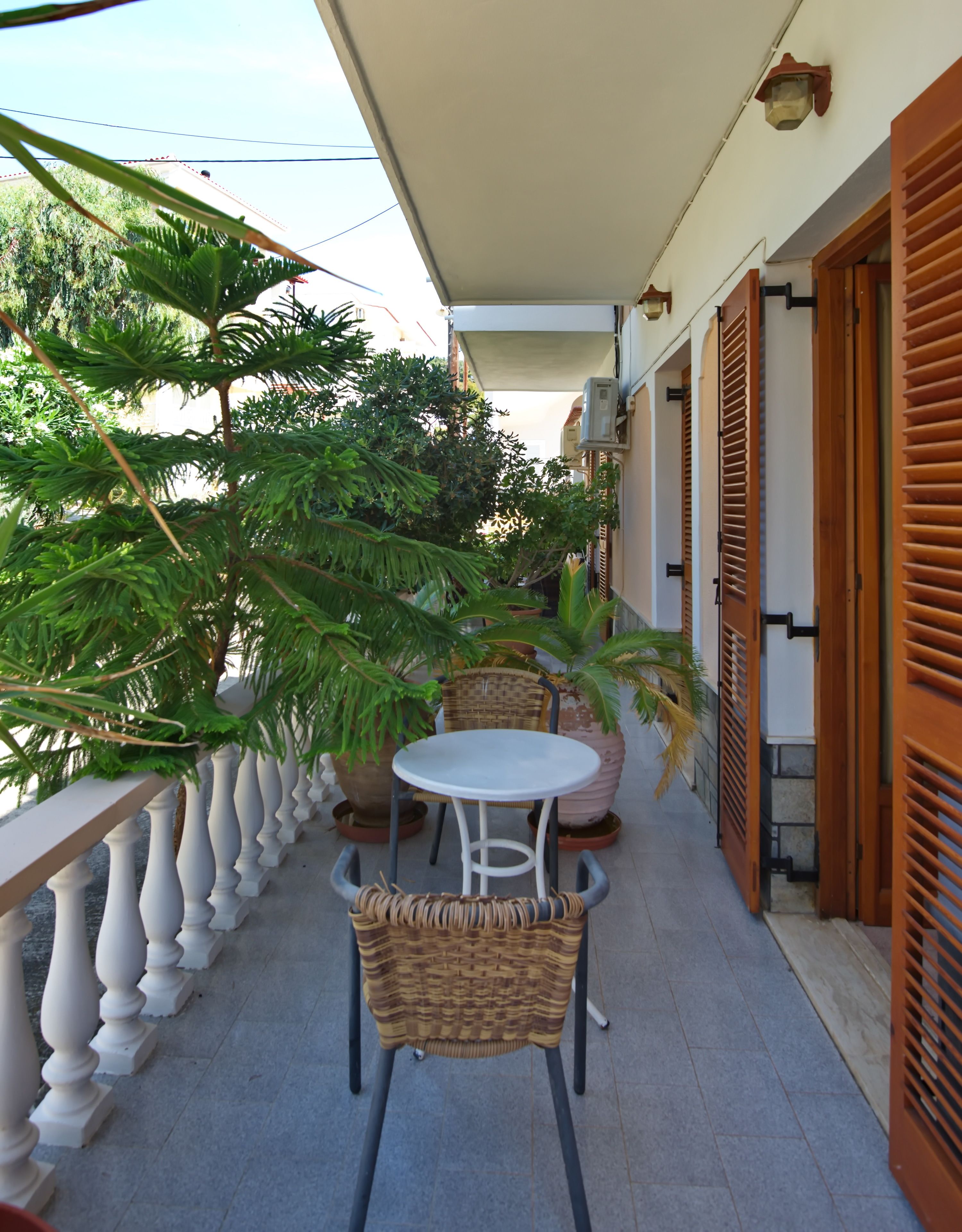 A balcony with a white table and wicker chairs, surrounded by potted plants and a railing.