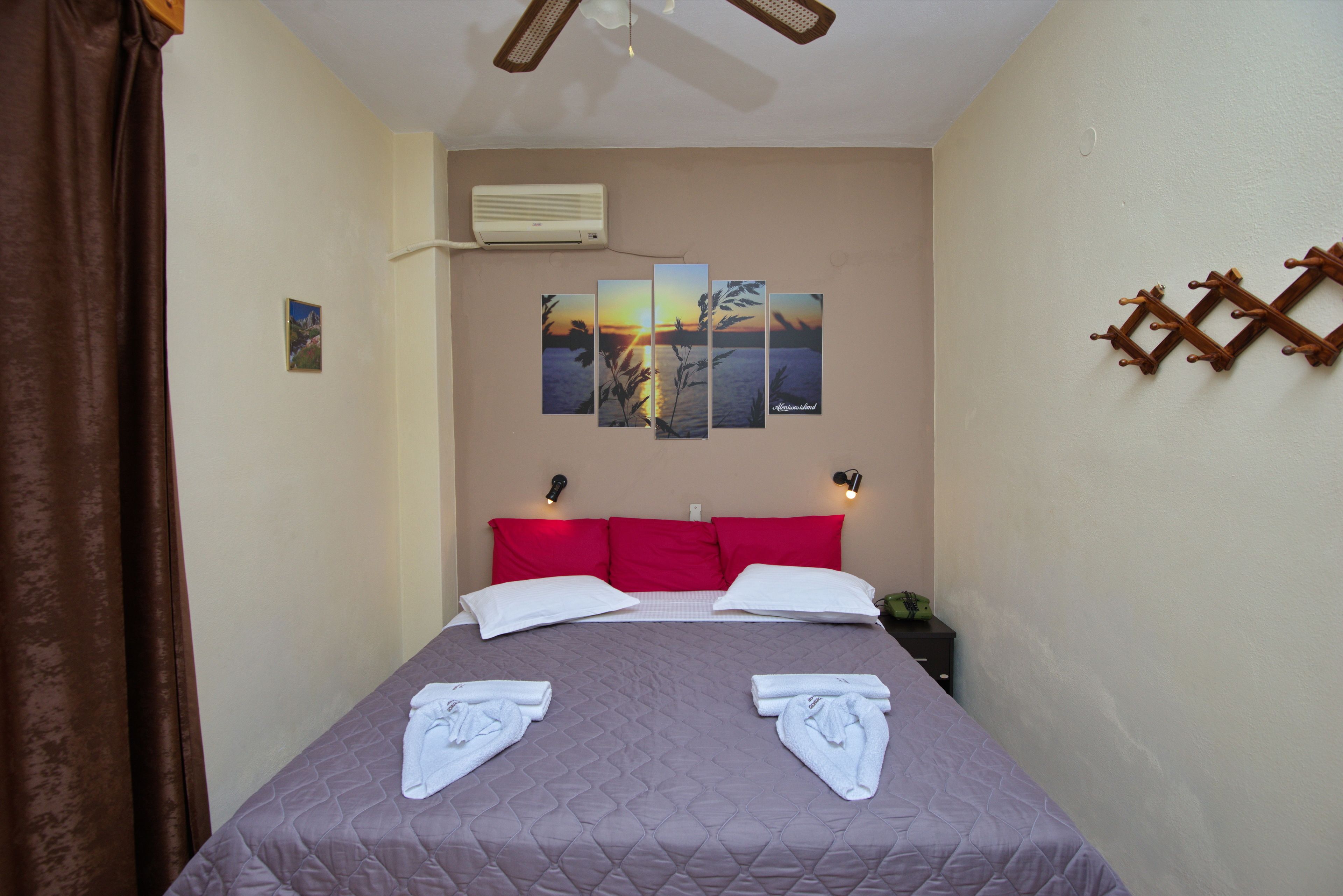 A hotel room with a bed, red pillows, a wall-mounted air conditioner, and a set of wall-mounted hooks.