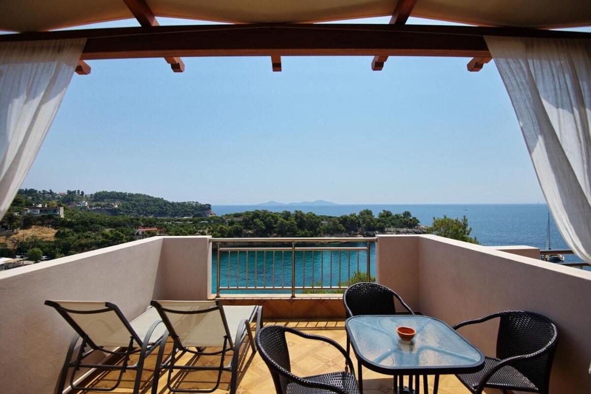 A balcony with a table and chairs, a view of the sea, and a distant landscape.