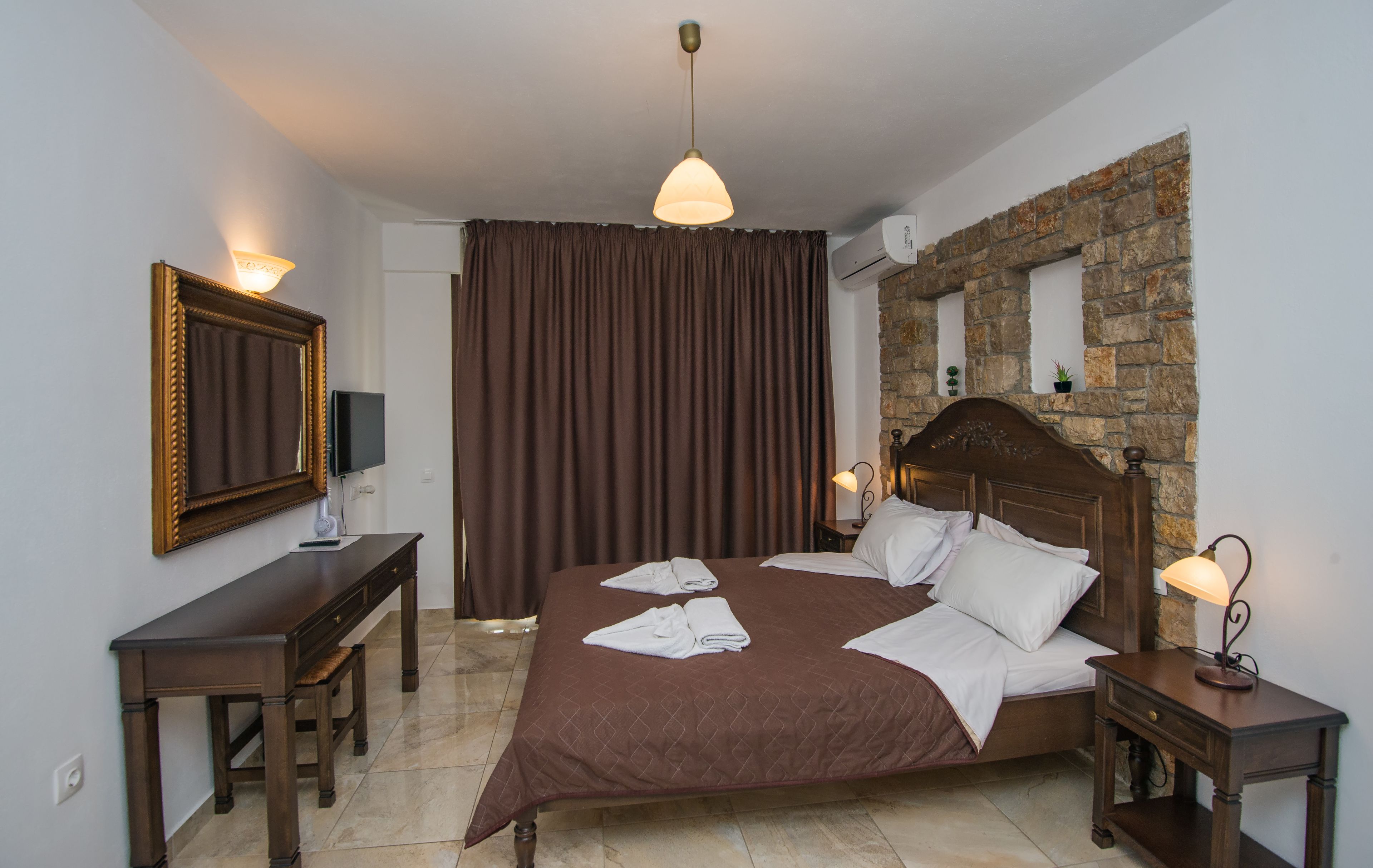 A hotel room with a large bed, a wooden nightstand, a desk, a mirror, and a stone accent wall.