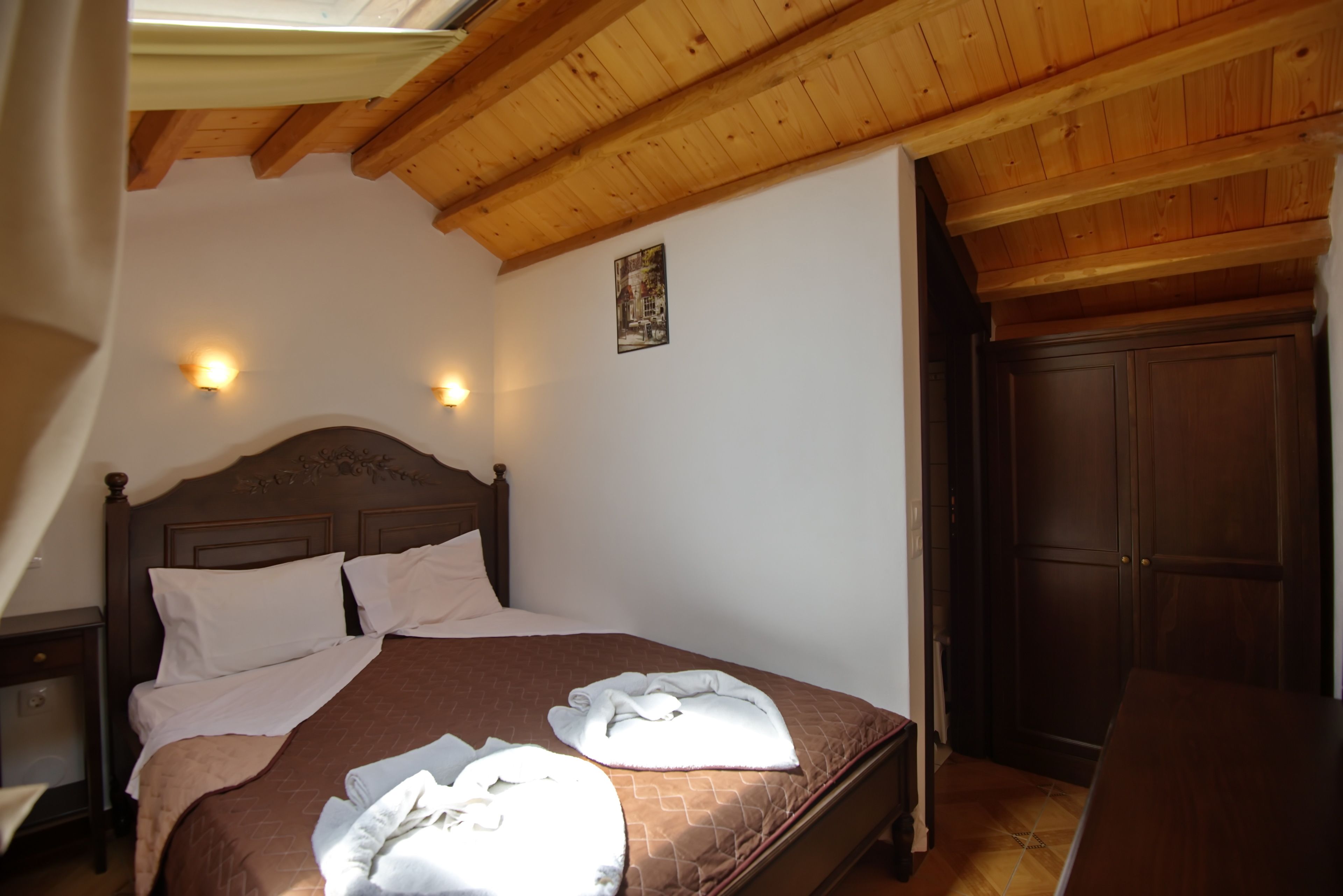 A bedroom with a wooden ceiling, a bed with a brown quilt, two white towels, and a nightstand.