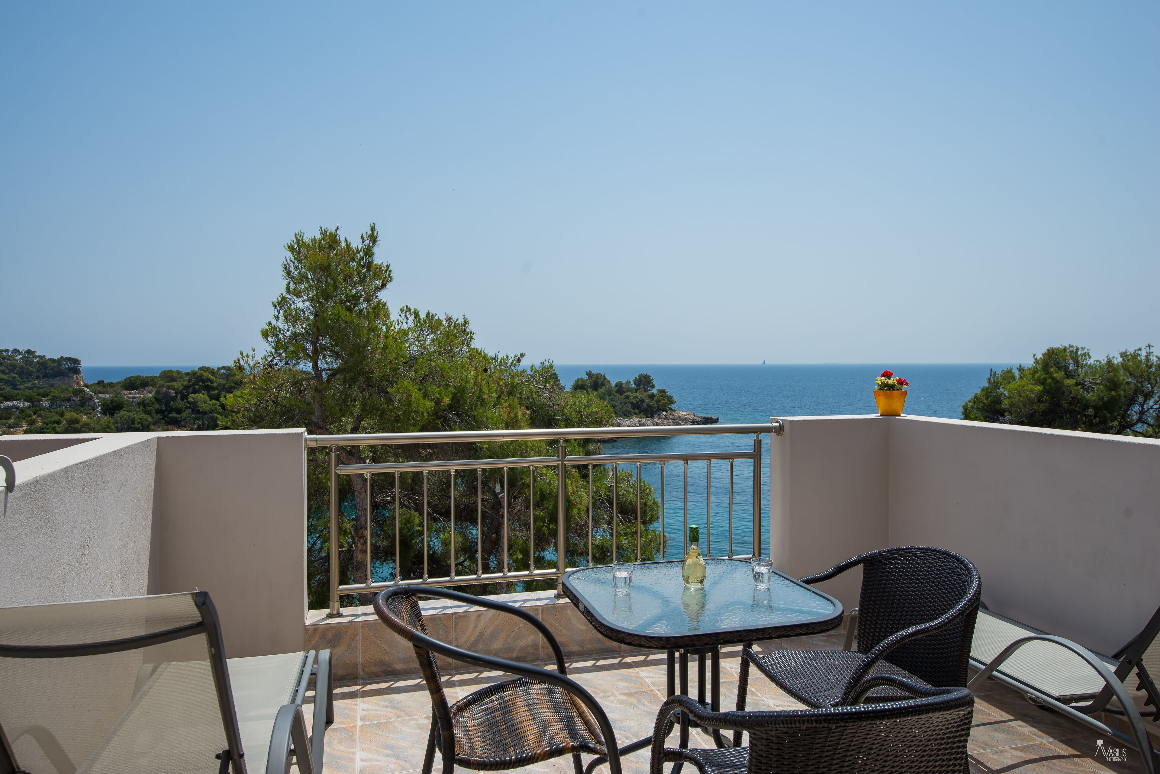 A balcony with a table and chairs, overlooking a scenic view of the sea and greenery.