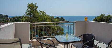 A balcony with a table and chairs, overlooking a scenic view of the sea and greenery.