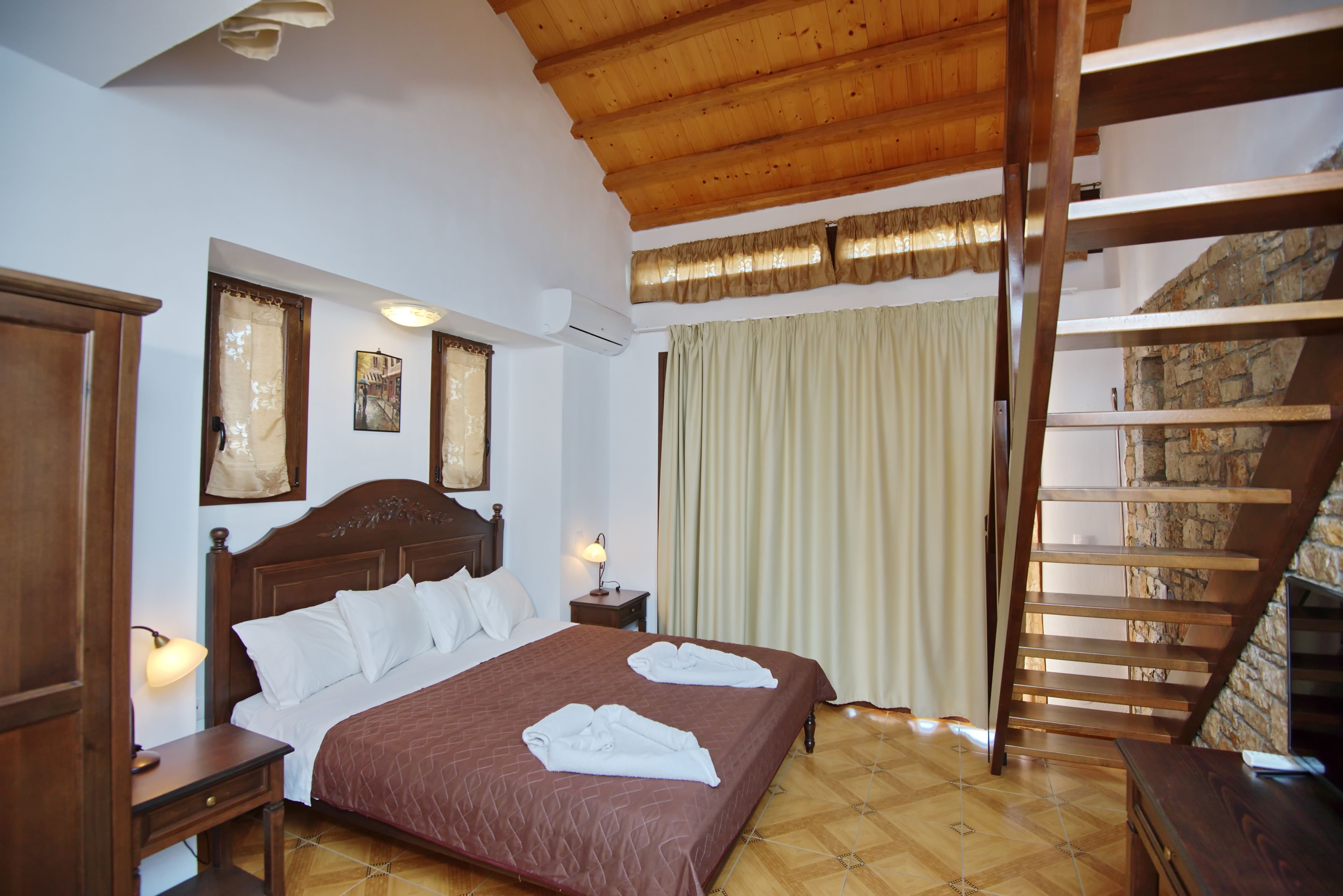A bedroom with a wooden bed, a nightstand, a lamp, a window with curtains, and a wooden staircase.