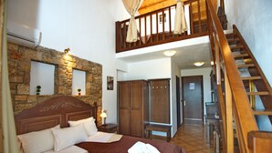 Quarto com cabeceira de madeira, uma cama grande com roupa de cama branca, um banco de madeira e uma escada que leva a um andar superior.