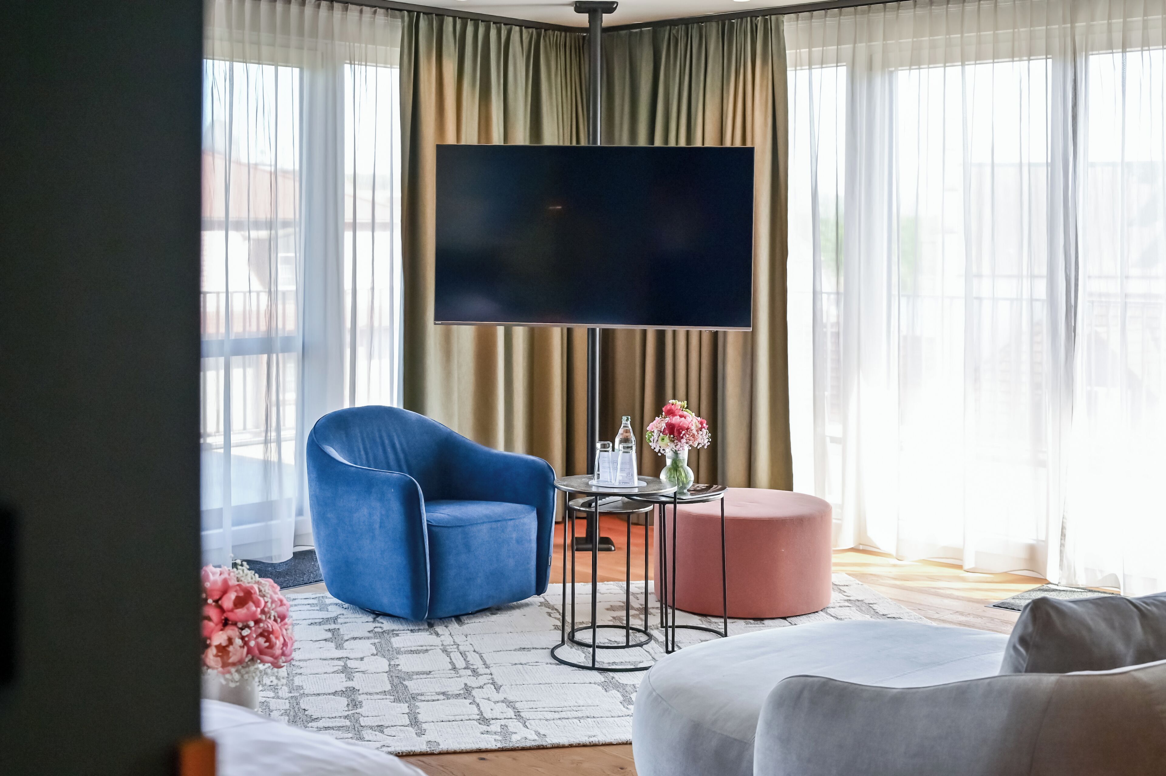 A hotel room with a large flat-screen TV, a blue armchair, a small round table with a vase of flowers, and a pink ottoman.