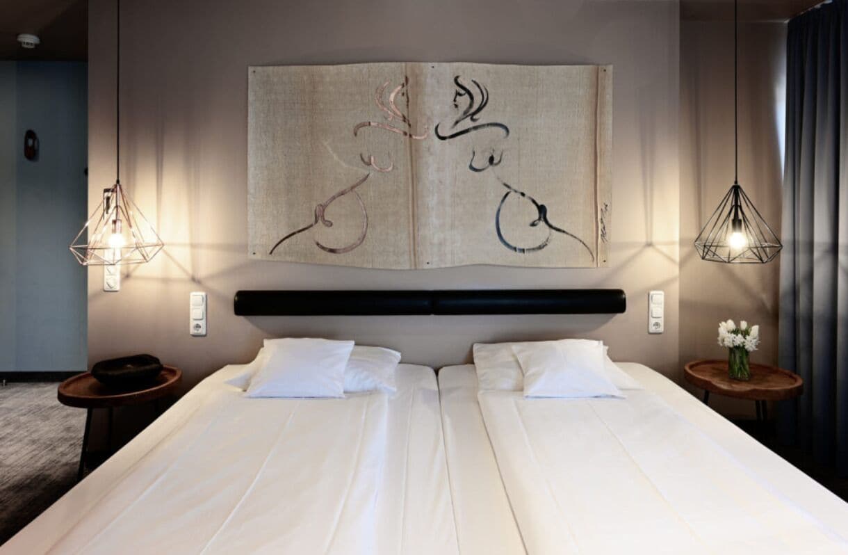 A hotel room with a large bed, two bedside tables, a wall-mounted artwork, and pendant lights.