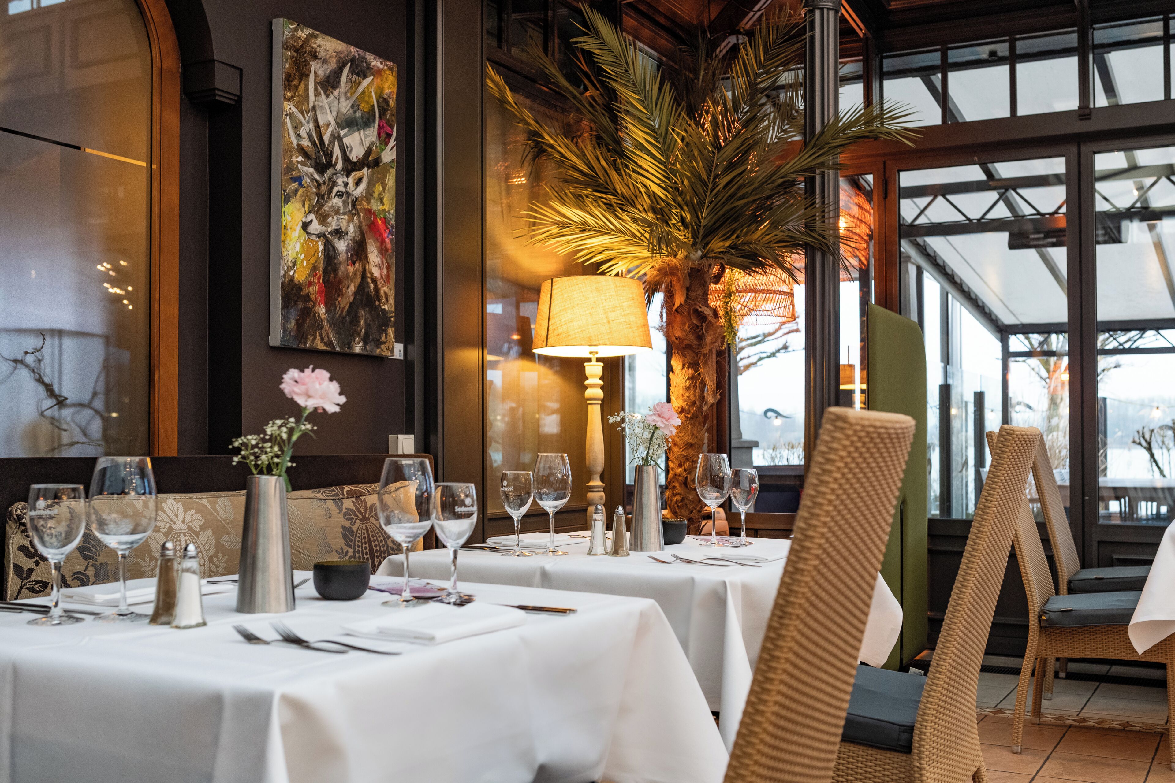 A restaurant interior with tables set for dining, a large painting on the wall, and a palm plant.