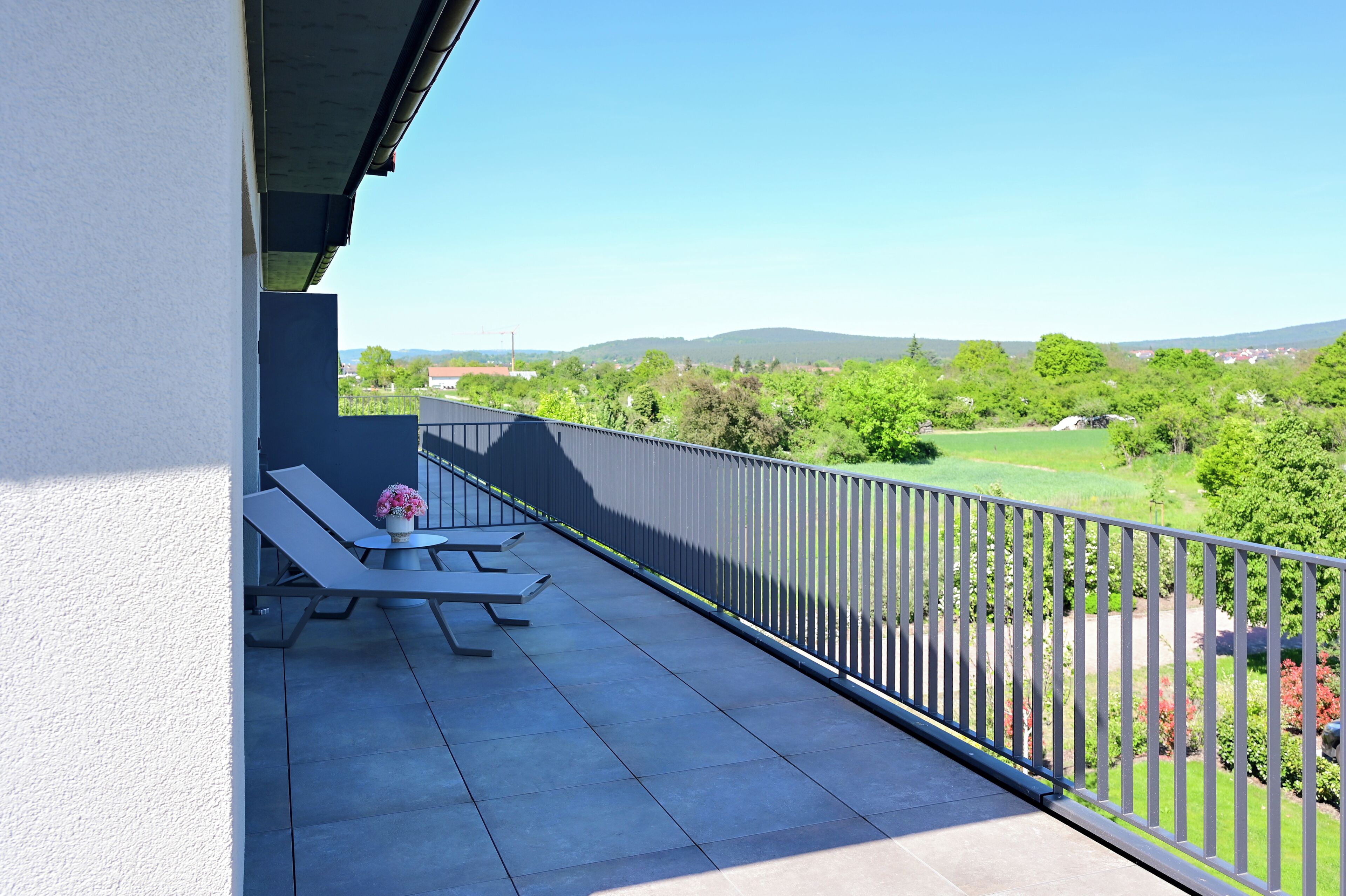 A balcony with two lounge chairs and a table, overlooking a green landscape with trees and distant hills.