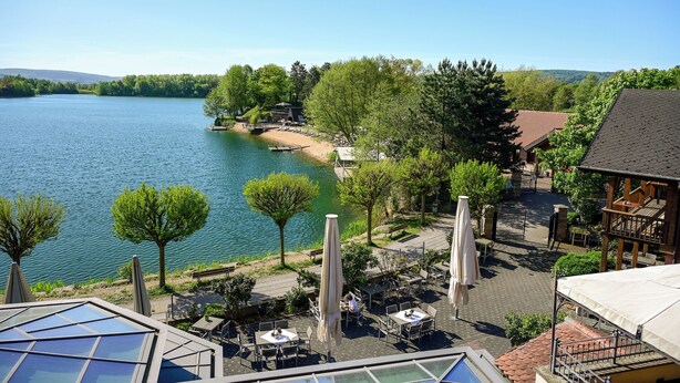 Vista de un lago con mobiliario exterior, un muelle para botes y un edificio con techo inclinado.