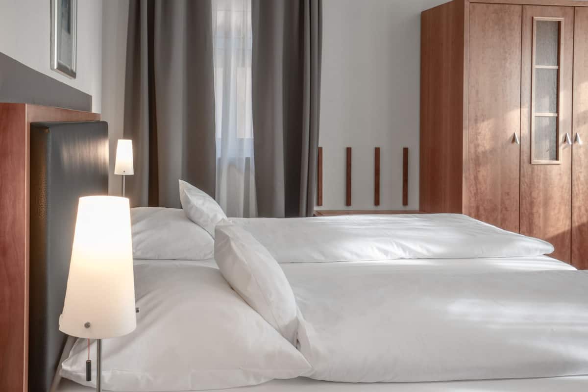Una habitación de hotel con cama, mesita de noche, lámpara, armario y ventana con cortinas.