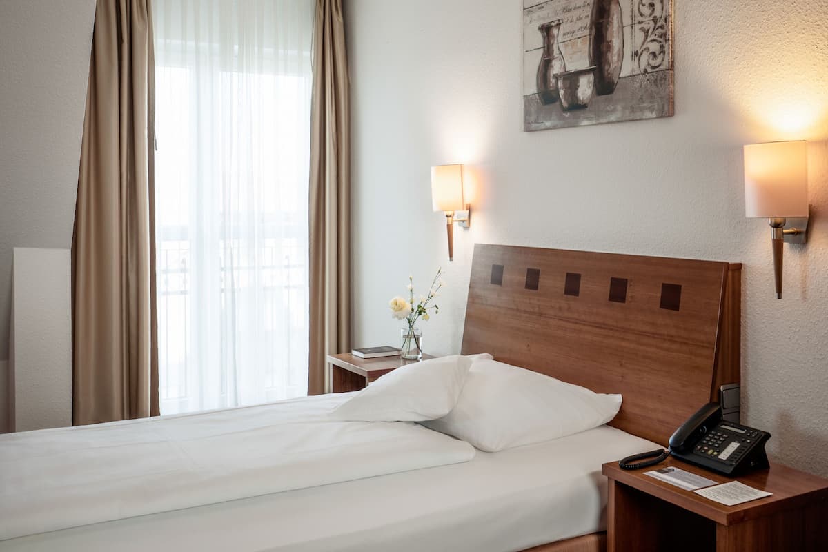 Habitación de hotel con cabecera de madera, cama con ropa de cama blanca, mesita de noche con lámpara, teléfono y un jarrón con flores.