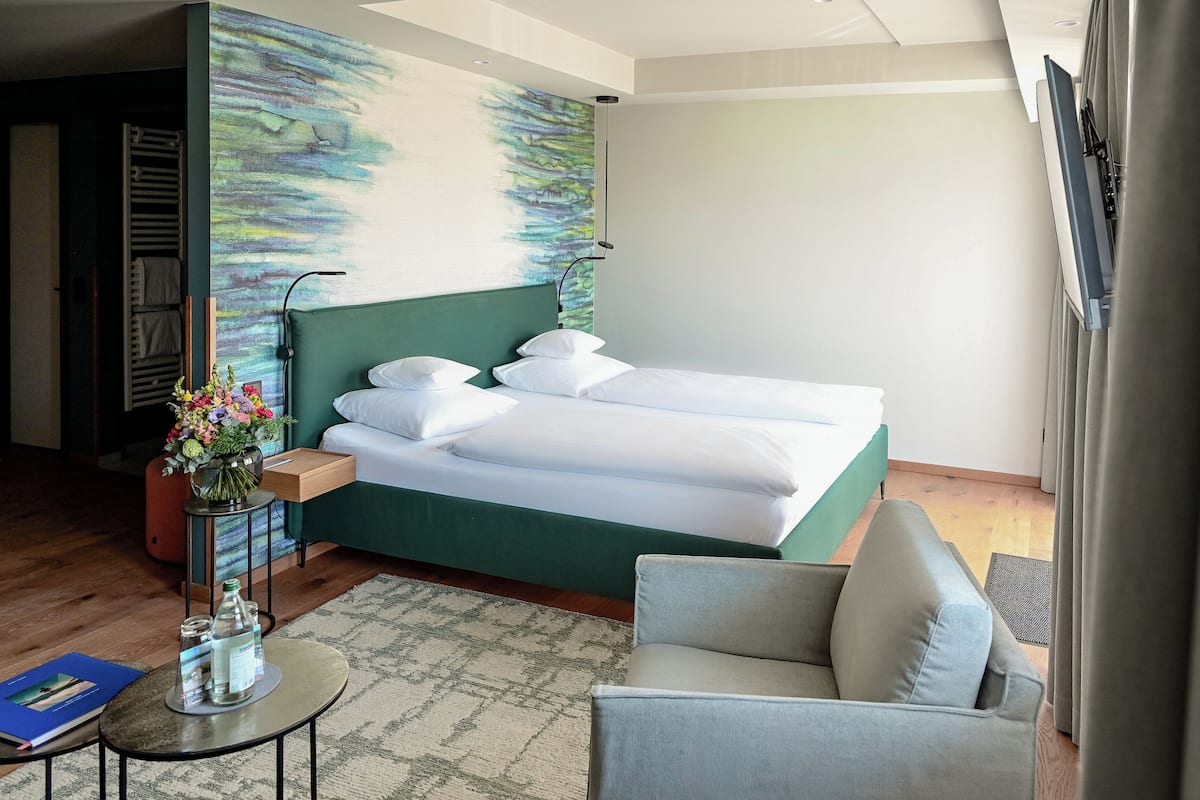 Habitación de hotel con una cama, una silla, una mesita con un florero y un cuadro en la pared.
